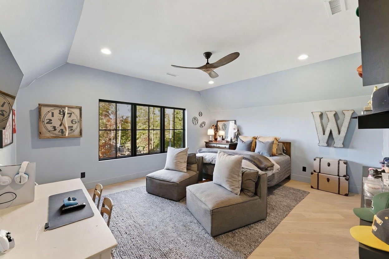 This spacious bedroom features light blue walls, a vaulted ceiling with a modern ceiling fan, and light-toned wood flooring. The room is furnished with a bed, two plush gray armchairs, and a desk area, all arranged on a large textured area rug. Large windows with black frames provide ample natural light and a view of the surrounding trees.