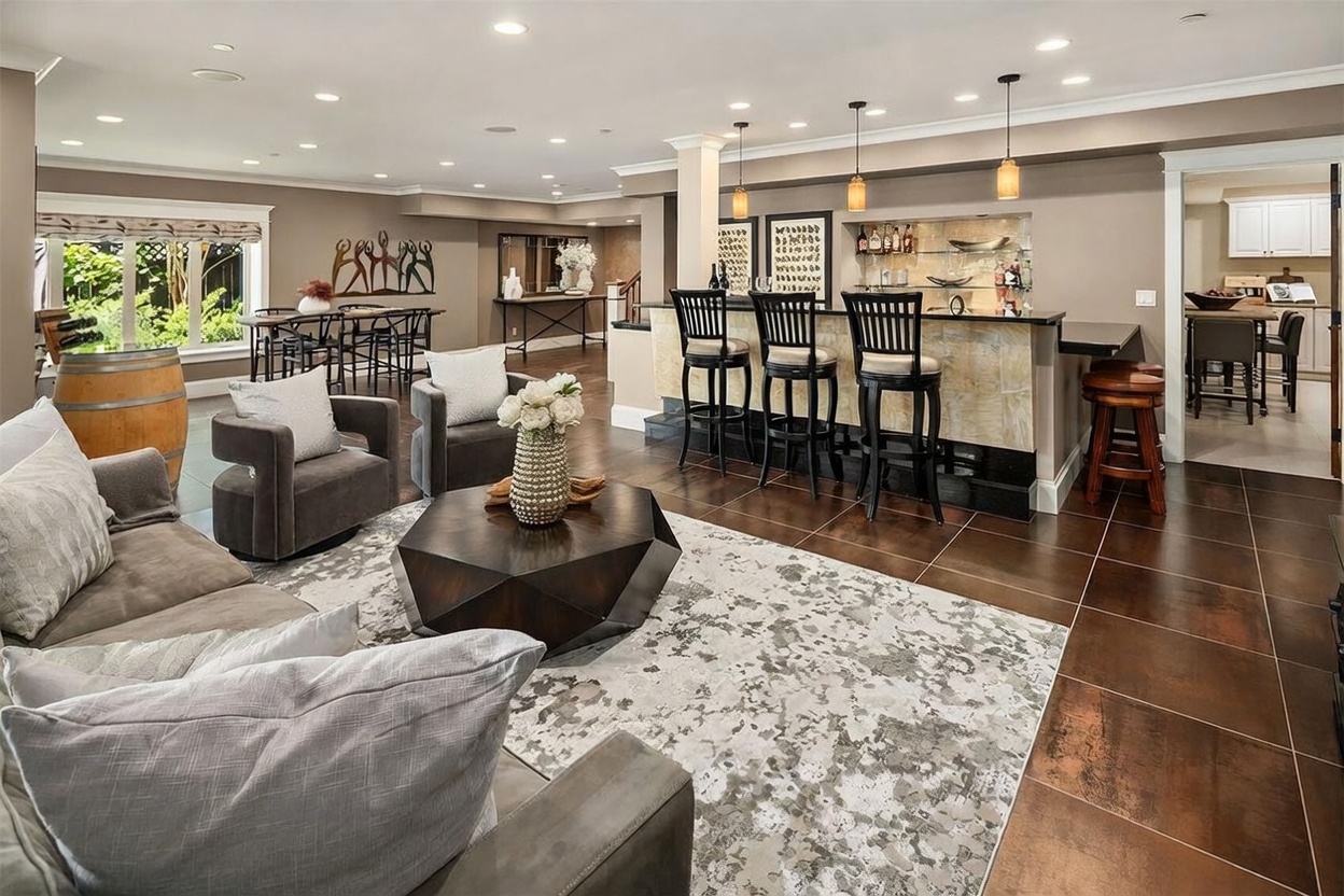 This spacious, open-concept basement features a sophisticated lounge area with a large, dark-toned sectional sofa and a geometric coffee table centered on a patterned area rug. A built-in bar with three high-backed stools serves as a focal point, illuminated by three pendant lights, while a separate dining area is visible in the background near a window. The room is finished with dark tile flooring and recessed lighting, creating a polished and inviting atmosphere for entertaining.