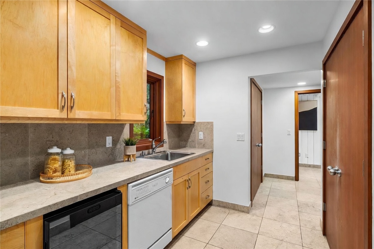 This kitchen features light wood cabinetry, stone-look countertops, and a neutral tile floor, creating a warm and functional workspace. The layout includes a built-in dishwasher, a stainless steel sink, and a small beverage cooler, all illuminated by recessed ceiling lighting. A patterned area rug adds a touch of texture to the space, which leads into a hallway, suggesting a well-connected home floor plan.