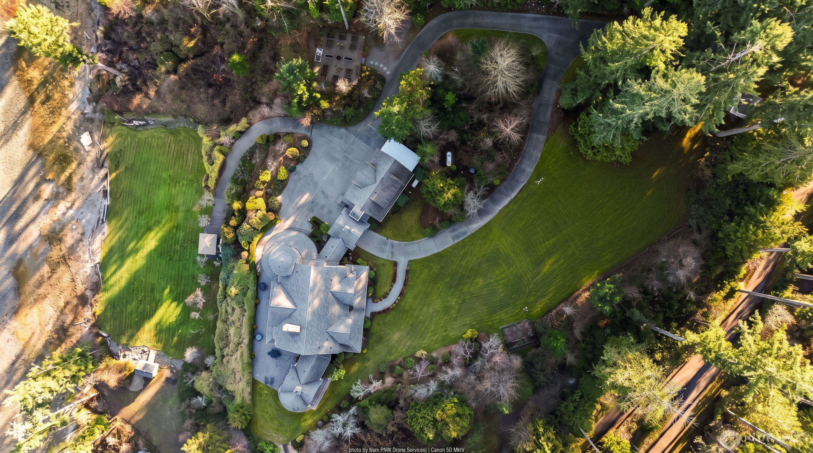 This high-angle aerial perspective captures a sprawling estate nestled within a lush, wooded landscape. The property features a large residence with a complex roofline, a private tennis court, and extensive paved driveways winding through mature trees and manicured lawns. The setting offers a sense of seclusion and luxury, highlighting the expansive grounds and outdoor recreational amenities.