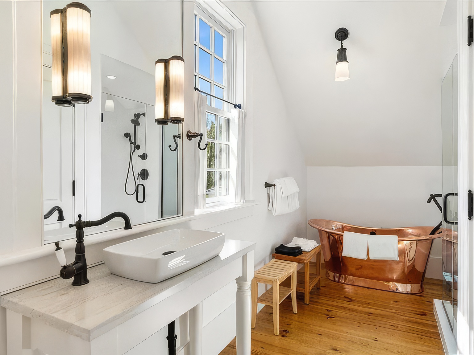 This bright and airy bathroom features a striking copper soaking tub as its centerpiece, set against a backdrop of white walls and warm wood flooring. A sleek vessel sink sits atop a white vanity with a stone countertop, complemented by dark bronze fixtures and wall-mounted lighting. The perspective captures the room from the doorway, showcasing the elegant contrast between the metallic tub and the clean, classic design elements.