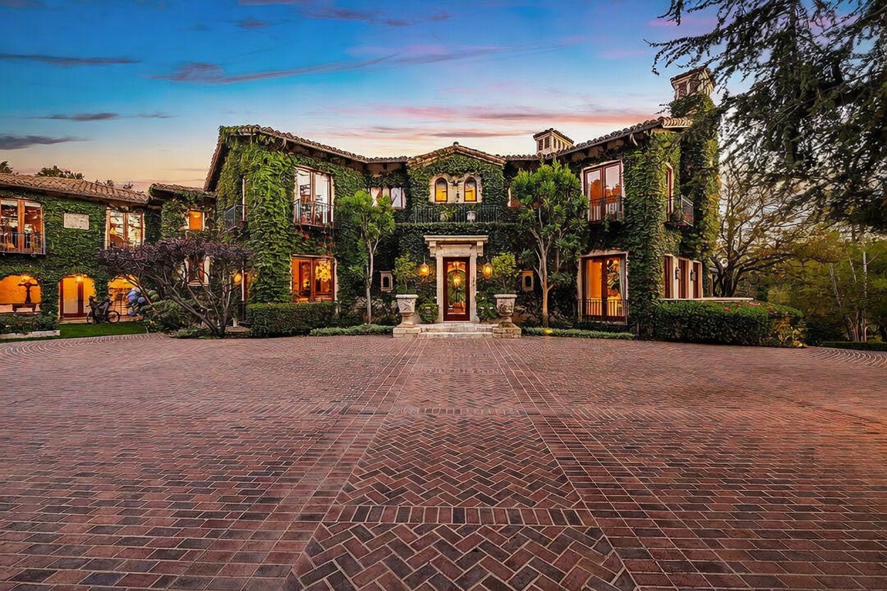 This grand, ivy-covered estate features a classic Mediterranean architectural style with a symmetrical facade and a prominent central entryway. A sprawling herringbone-patterned brick driveway leads directly to the front door, framed by mature trees and lush landscaping. The warm, golden hour lighting enhances the rich textures of the stone walls and the intricate ironwork on the balconies.
