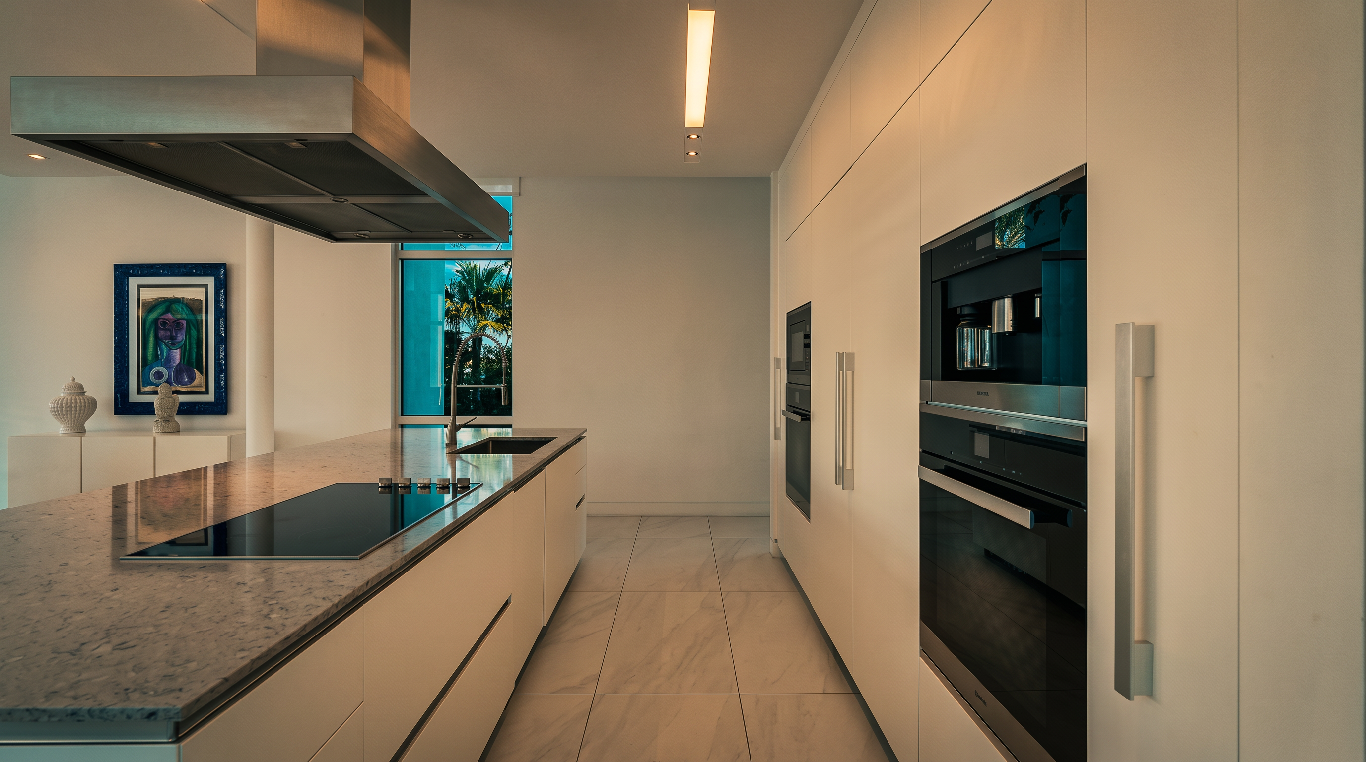 This sleek, modern kitchen features minimalist white cabinetry and a large central island topped with a light-toned stone surface. A stainless steel range hood hangs above the island, while built-in appliances are integrated into the wall cabinetry. The space is brightened by recessed lighting and large windows that provide a view of lush greenery outside.