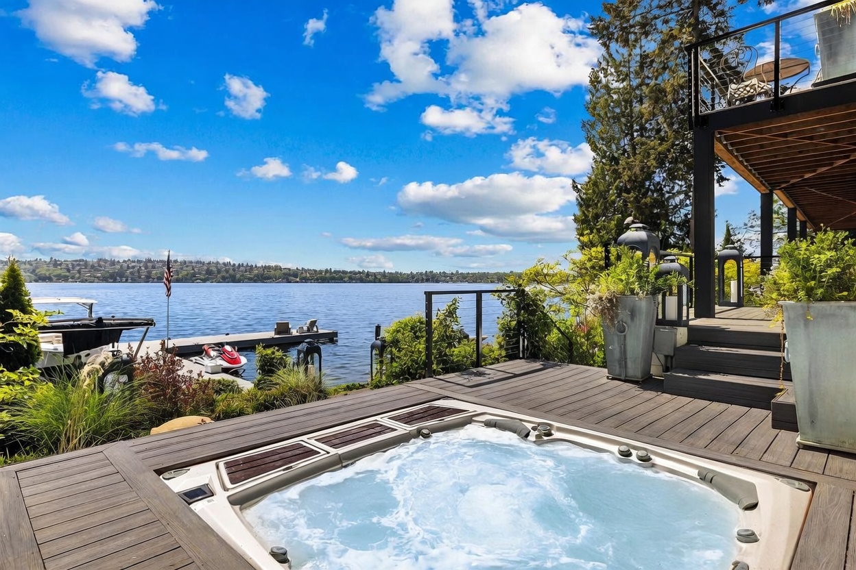 This elevated deck features a built-in hot tub as the central focal point, overlooking a serene lake with a private dock and a jet ski. The wooden decking provides a warm, natural aesthetic, while stairs lead up to a secondary balcony level above. The expansive water view and clear blue sky create a tranquil, resort-like atmosphere perfect for outdoor relaxation.