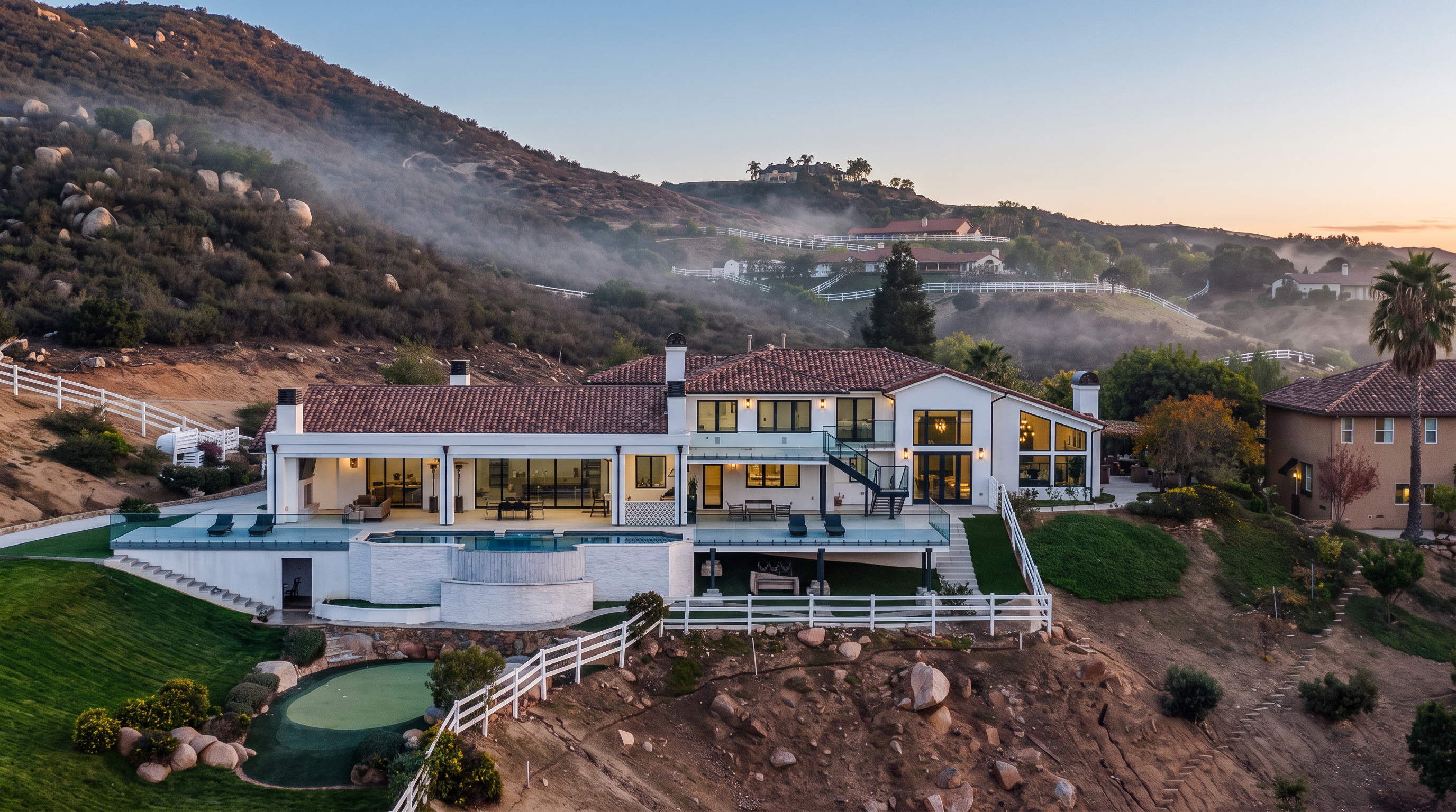This stunning Mediterranean-style estate features an expansive rear view, showcasing a multi-level layout with a prominent patio area, a swimming pool, and a private putting green. The home boasts large floor-to-ceiling windows and sliding glass doors that seamlessly integrate indoor and outdoor living against a dramatic sunset backdrop. Its elevated position on a hillside offers a commanding presence and panoramic views of the surrounding mountainous landscape.