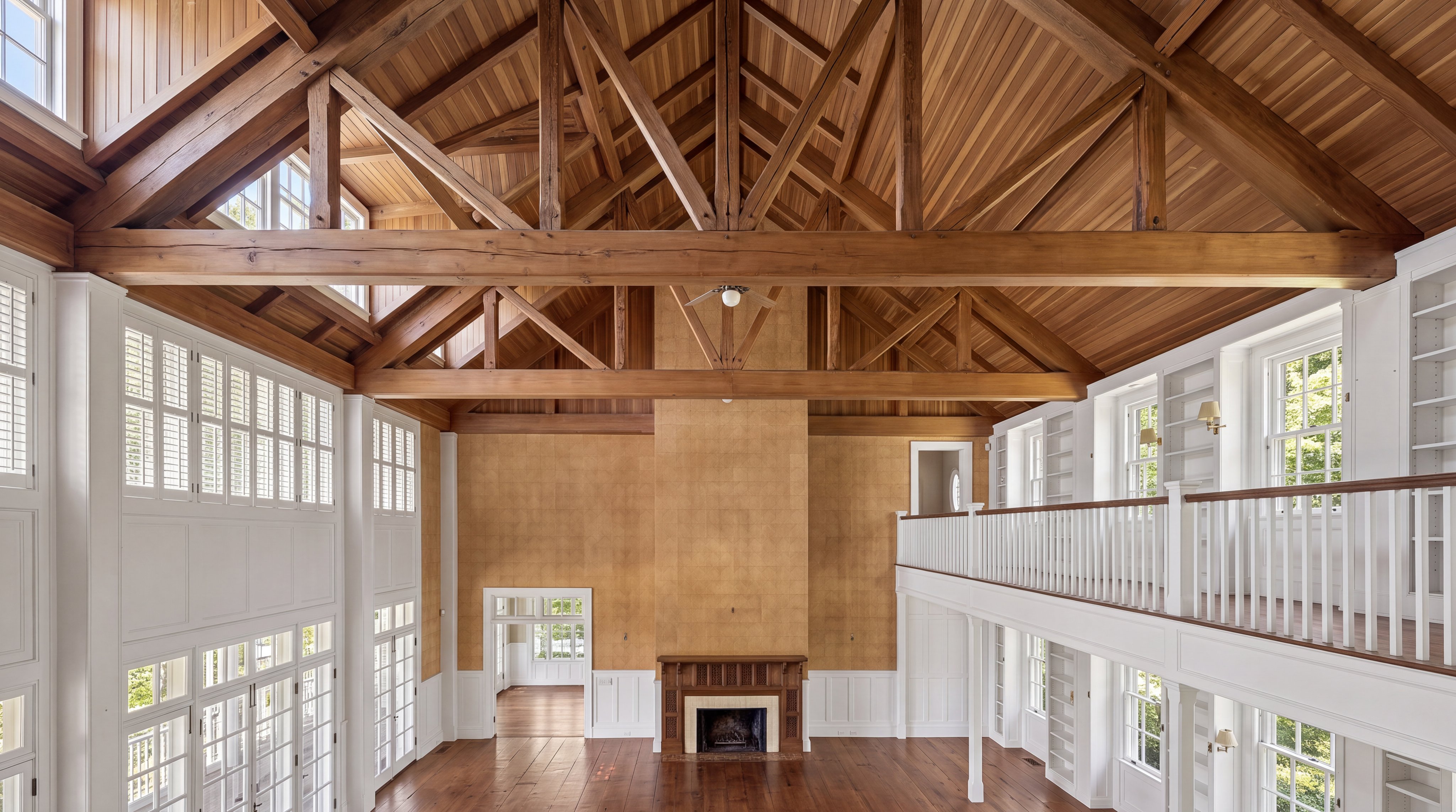 This grand living room features a double-height vaulted ceiling with exposed wooden trusses and beautiful wood paneling. A large, central fireplace serves as the focal point, complemented by a mezzanine balcony, built-in shelving, and floor-to-ceiling windows that provide an abundance of natural light. The rich wood floors and intricate architectural details create a warm, sophisticated, and spacious atmosphere.