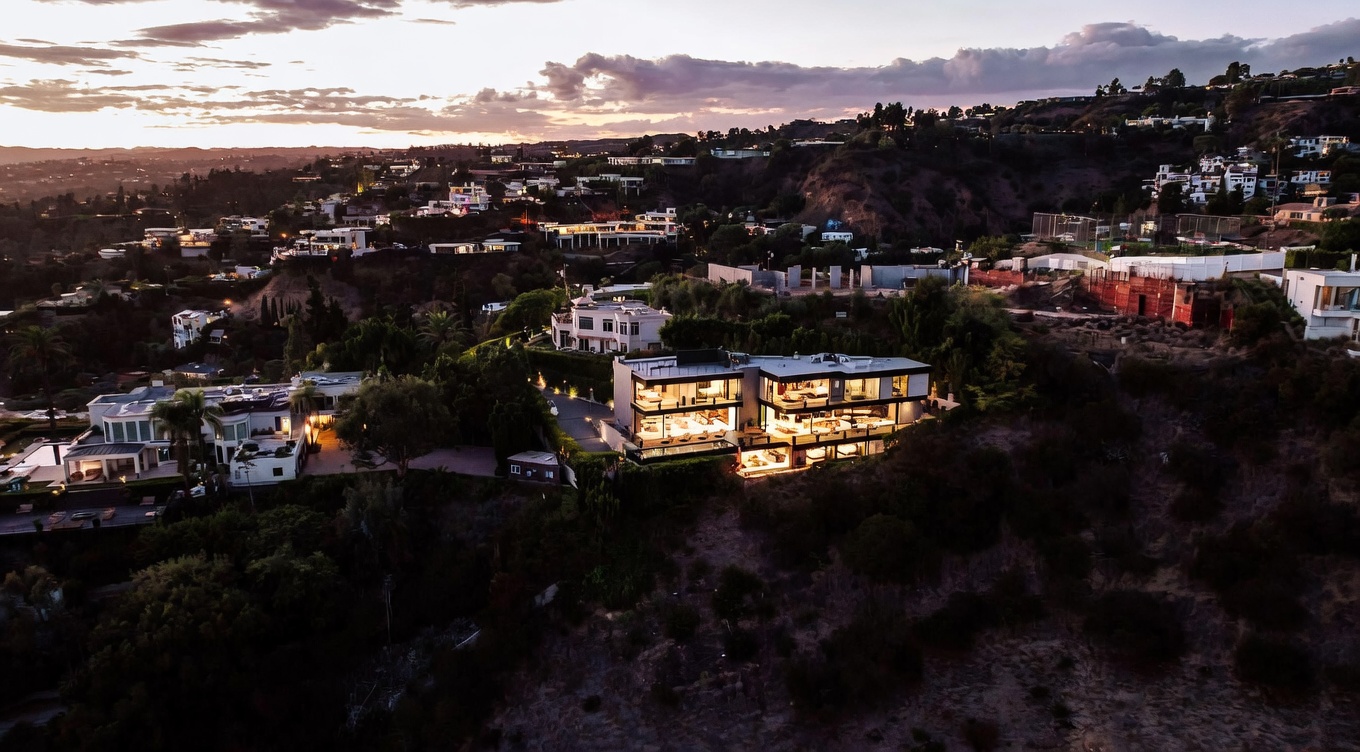 This stunning aerial perspective captures a luxurious, multi-level modern mansion perched on a hillside during a dramatic sunset. The home features expansive glass walls, multiple illuminated terraces, and a sleek architectural design that integrates seamlessly with the rugged landscape. The surrounding neighborhood is nestled into the rolling hills, creating an atmosphere of exclusivity and high-end living.