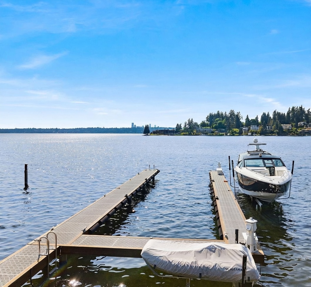 This image captures a serene waterfront view from a private dock extending into a large, calm lake. A sleek motorboat is secured on a lift to the right, while a smaller covered vessel rests at the end of the dock, emphasizing the property's recreational appeal. In the distance, the faint silhouette of a city skyline is visible across the water, framed by lush green trees along the shoreline under a bright, clear blue sky.