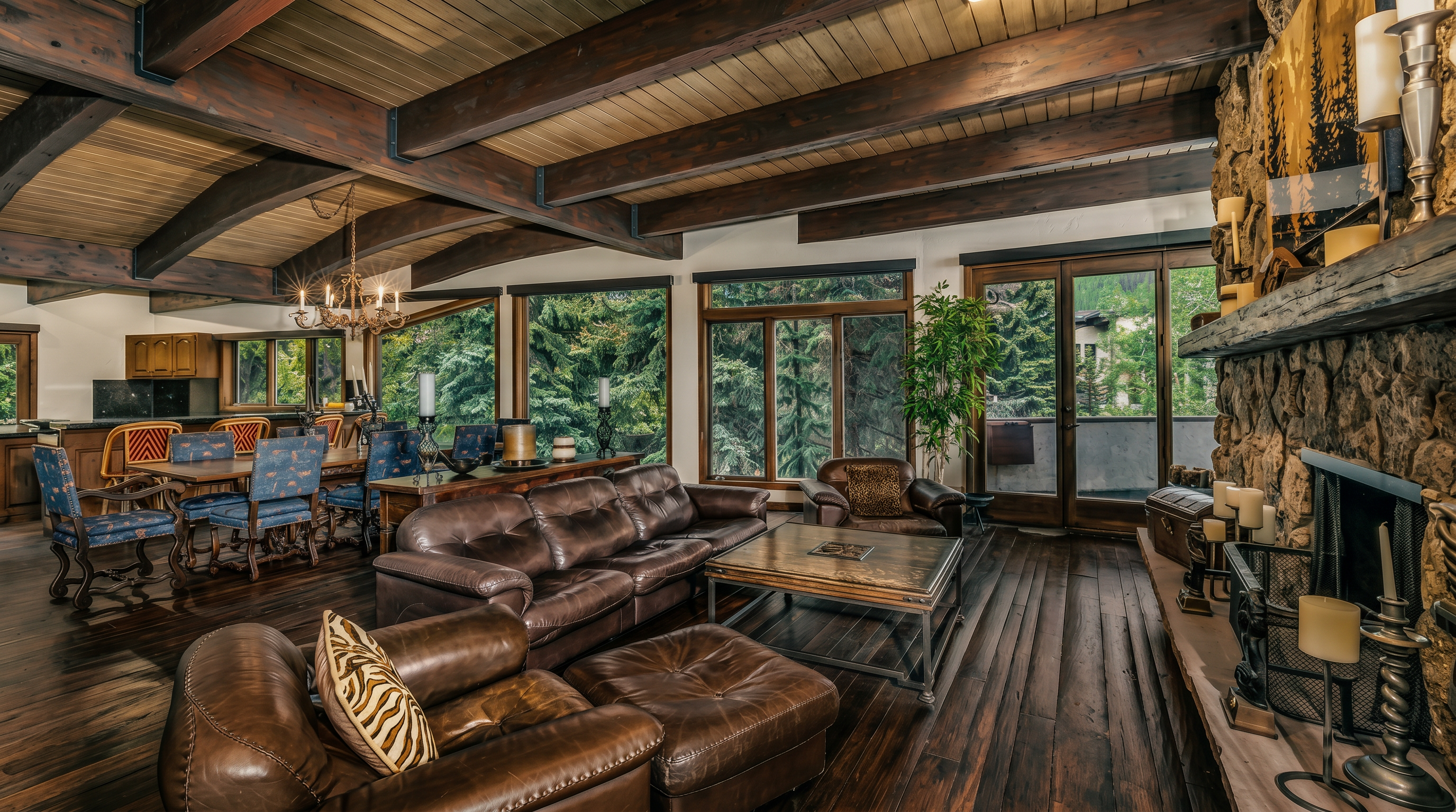 This spacious living room features a rustic aesthetic with exposed wooden ceiling beams and dark wood flooring. A large stone-clad chimney breast serves as a focal point, complemented by a leather sofa set and a central coffee table. Large windows provide ample natural light and views of the surrounding forest, creating a warm and inviting mountain-lodge atmosphere.
