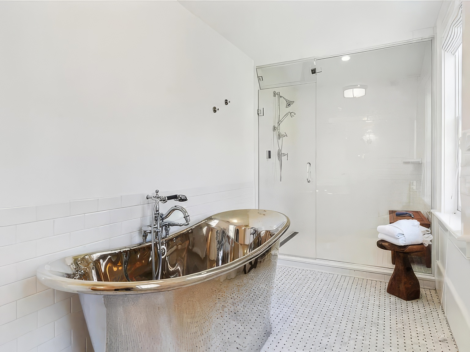 This elegant bathroom features a striking metallic, freestanding soaking tub as its centerpiece, set against a wall of white subway tile. To the right, a glass-enclosed walk-in shower with multiple showerheads offers a modern, spa-like experience. The space is finished with a classic patterned tile floor and a rustic wooden stool, creating a sophisticated blend of contemporary and organic textures.