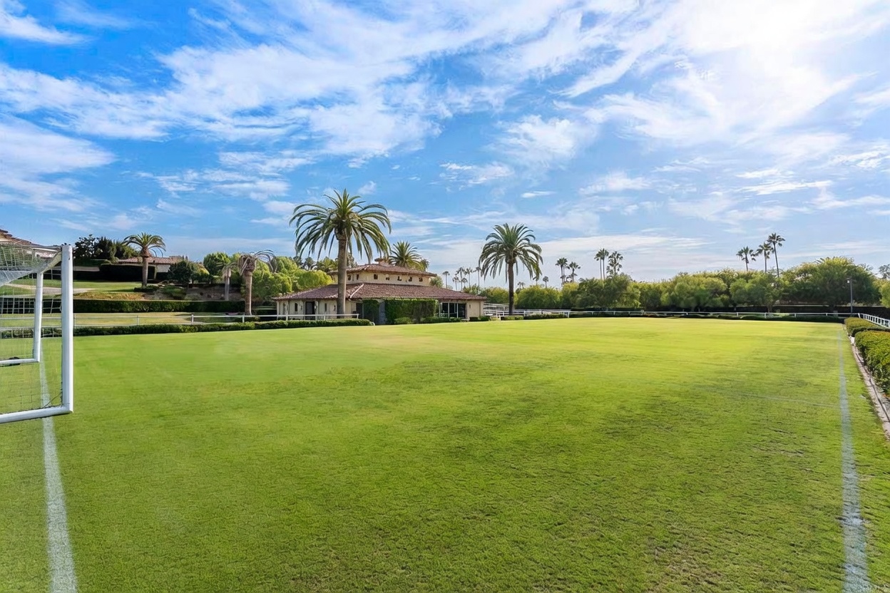 This image showcases a vast, meticulously maintained lawn, possibly a sports field, with soccer goals visible on either side. In the background, a luxurious building with a red tile roof is nestled among palm trees and lush greenery, suggesting a high-end estate. The clear blue sky with scattered clouds enhances the overall impression of spaciousness and tranquility.