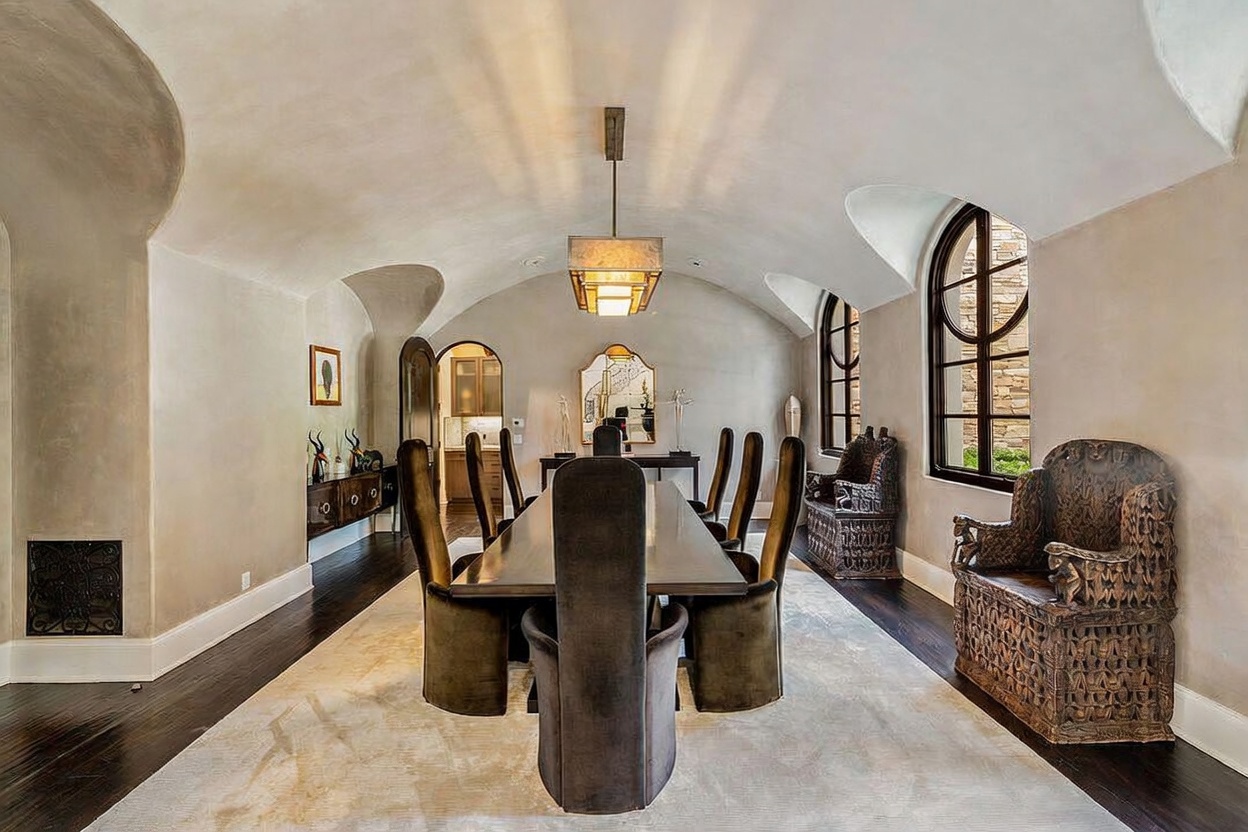 This formal dining room features a dramatic barrel-vaulted ceiling and dark hardwood flooring, creating an elegant and sophisticated atmosphere. A long, dark-toned dining table is surrounded by high-backed upholstered chairs, centered beneath a large, modern rectangular pendant light. The room is accented by ornate, carved wooden chairs placed against the side walls and a decorative mirror reflecting the space, all framed by arched windows that allow natural light to enter.