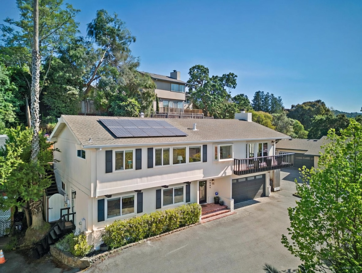 This elevated front-view perspective showcases a two-story suburban home with a light-colored exterior, dark shutters, and a prominent solar panel array on the roof. The property features a paved driveway leading to a garage, a small balcony off the upper level, and a neatly manicured hedge lining the front foundation. The setting is bright and sunny, framed by mature trees and a clear blue sky, suggesting a peaceful residential neighborhood.