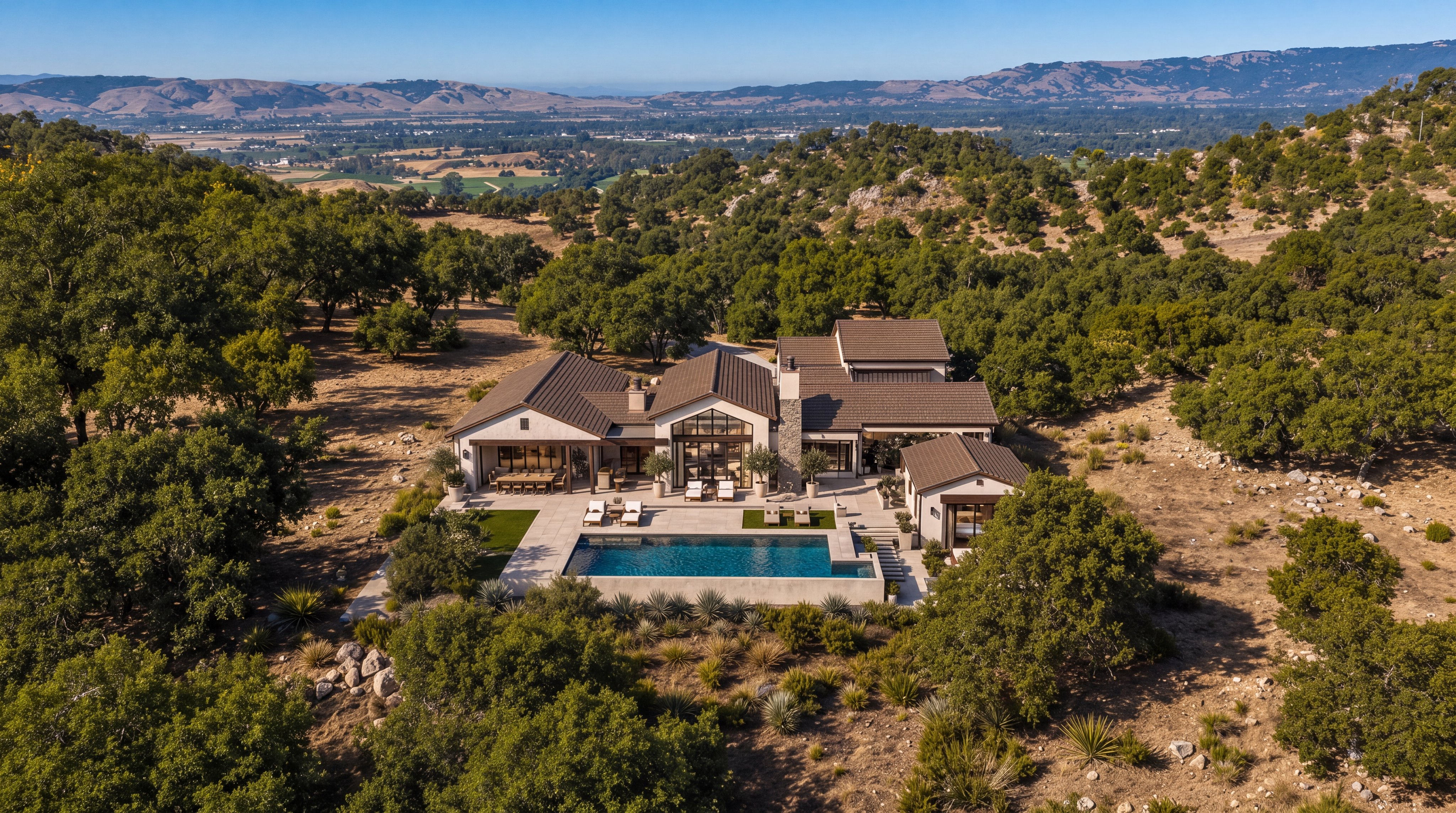 This stunning aerial view captures a contemporary farmhouse-style estate nestled in a scenic, rural landscape. The property features a spacious rear patio with a custom pool, surrounded by expansive, tree-dotted acreage that offers panoramic valley views. The home's neutral color palette and high-end design integrate harmoniously with the natural surroundings, creating a luxurious and serene retreat.