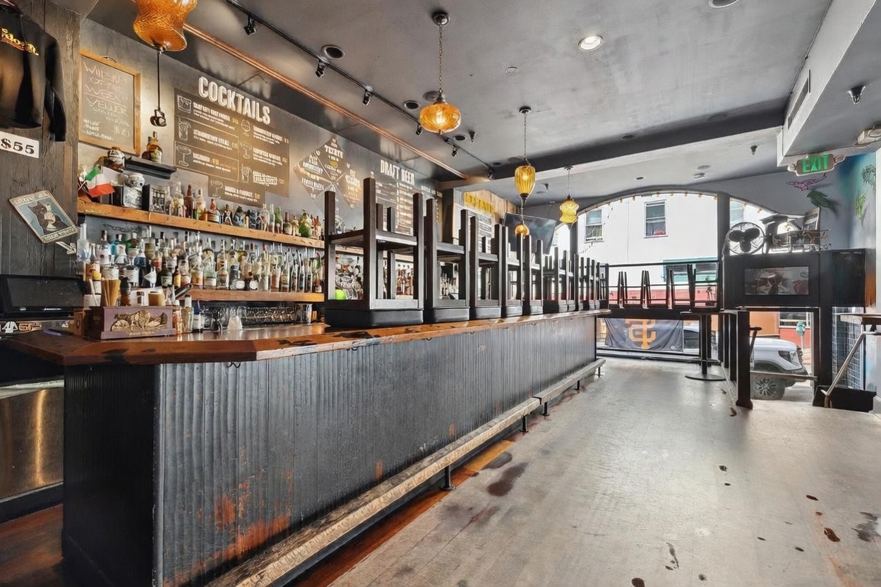 This image captures the interior of a commercial bar space, featuring a long, dark-toned wooden bar counter with chairs inverted on top for cleaning or closing. Behind the bar, a large chalkboard menu displays cocktail options, while shelves are stocked with an extensive collection of liquor bottles. The perspective is a wide-angle shot from the floor level, looking down the length of the bar toward a large arched window that allows natural light to enter the space.