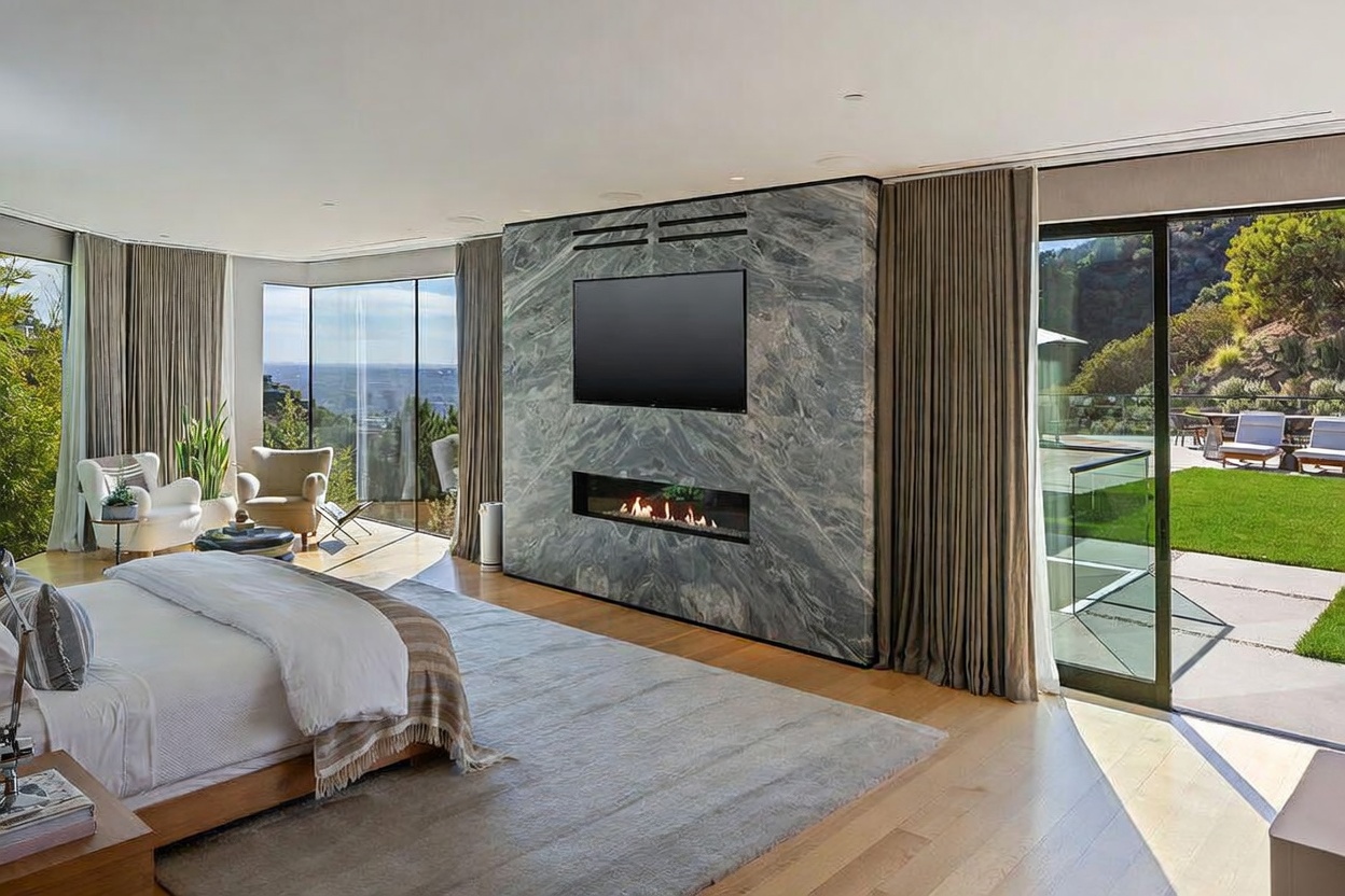This spacious primary bedroom features a prominent stone-clad chimney breast that serves as a focal point, housing both a television and a linear fireplace. The room is framed by expansive floor-to-ceiling glass walls that offer panoramic views and provide direct access to an outdoor patio area. Light wood flooring and a neutral color palette create a serene, modern atmosphere, complemented by soft drapery and comfortable seating near the windows.