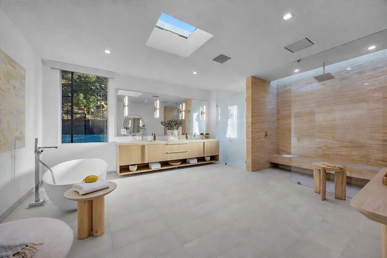 This expansive primary bathroom features a minimalist, spa-like aesthetic with light wood cabinetry, a freestanding soaking tub, and a large walk-in shower with stone-clad walls. A skylight above the vanity area floods the space with natural light, complementing the neutral color palette and clean lines. The perspective is wide-angle, capturing the entire room from the entrance to showcase the seamless integration of the vanity, bathing, and shower zones.