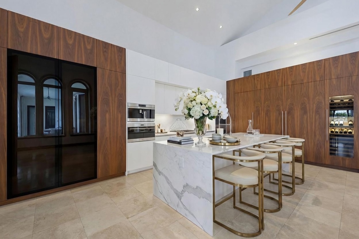 This modern kitchen features a large central island with a stone waterfall edge and integrated seating, complemented by sleek wood-paneled cabinetry and white upper cabinets. The space is equipped with high-end built-in appliances, including a wine cooler and double ovens, creating a sophisticated and functional culinary environment. The perspective is a wide-angle shot from the kitchen floor, emphasizing the clean lines and high-contrast design of the room.