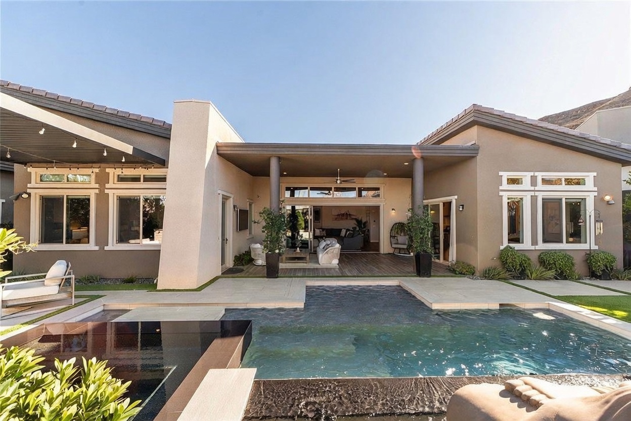 This rear view of a modern residence showcases a seamless transition between indoor and outdoor living spaces, featuring a covered patio that opens into a bright living area. A sleek, dark-tiled infinity-edge pool sits in the foreground, reflecting the clear sky and the home's neutral-toned stucco facade. The property is framed by manicured landscaping and a desert-inspired backdrop, creating a serene and luxurious atmosphere.