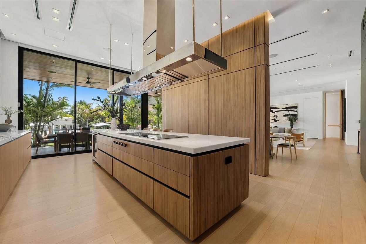 This modern, open-concept kitchen features a large central island with a stone countertop and light wood cabinetry. A sleek, stainless steel range hood hangs above the island, while floor-to-ceiling glass walls provide a seamless transition to the outdoor patio area. The space is bright and airy, with light wood flooring extending into the adjacent living and dining areas, creating a cohesive and sophisticated atmosphere.