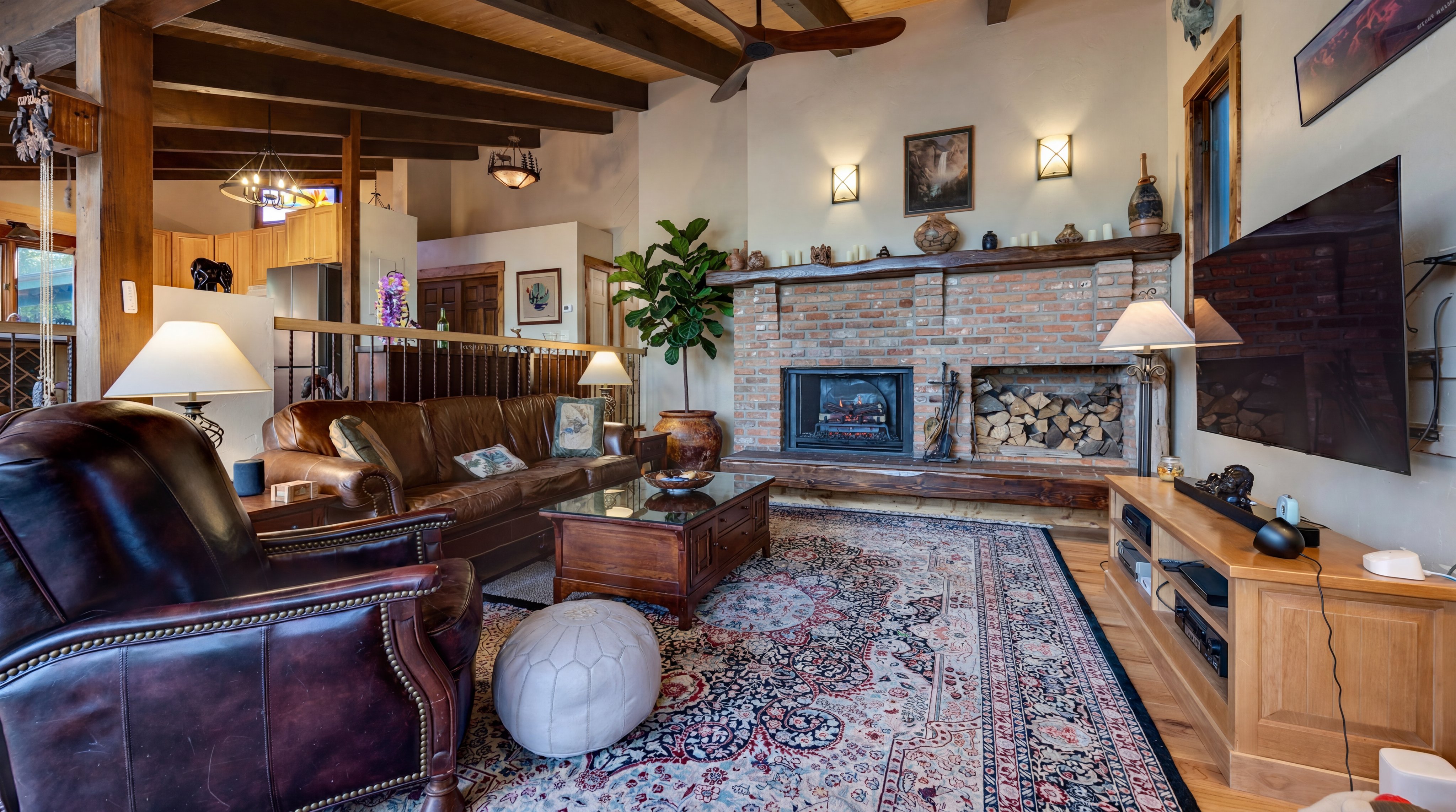 This cozy living room features a warm, rustic aesthetic with a prominent brick fireplace, vaulted wood-beam ceilings, and rich leather furniture. The inviting space is centered around a traditional patterned area rug and a wooden coffee table, offering a comfortable atmosphere accented by southwestern decor. A large flat-screen television and media console are integrated into the room, making it both a relaxing retreat and a functional entertainment area.