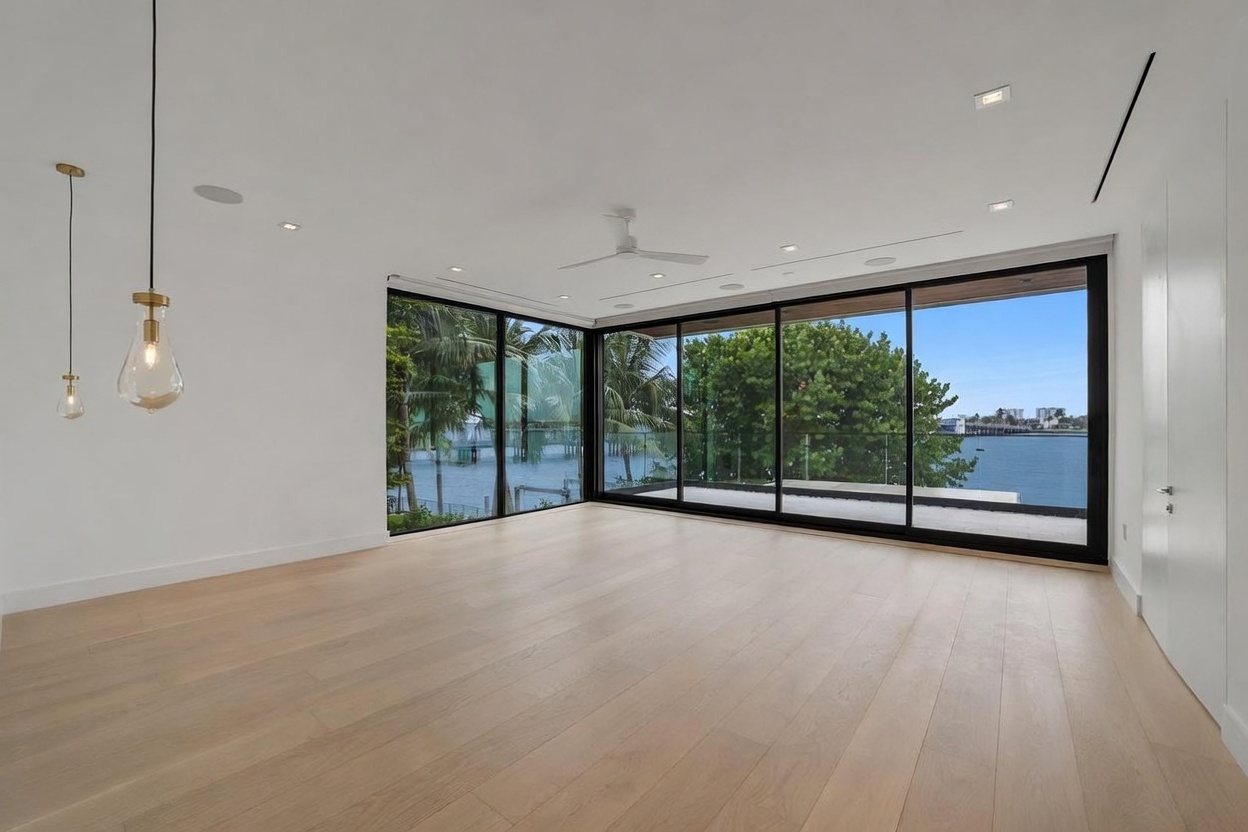 This bright, minimalist living space features light wood flooring and crisp white walls, creating an airy and open atmosphere. The room is defined by expansive floor-to-ceiling glass walls that provide panoramic views of the water and surrounding greenery. A modern ceiling fan and sleek pendant lighting fixtures add functional elegance to the clean, contemporary design.