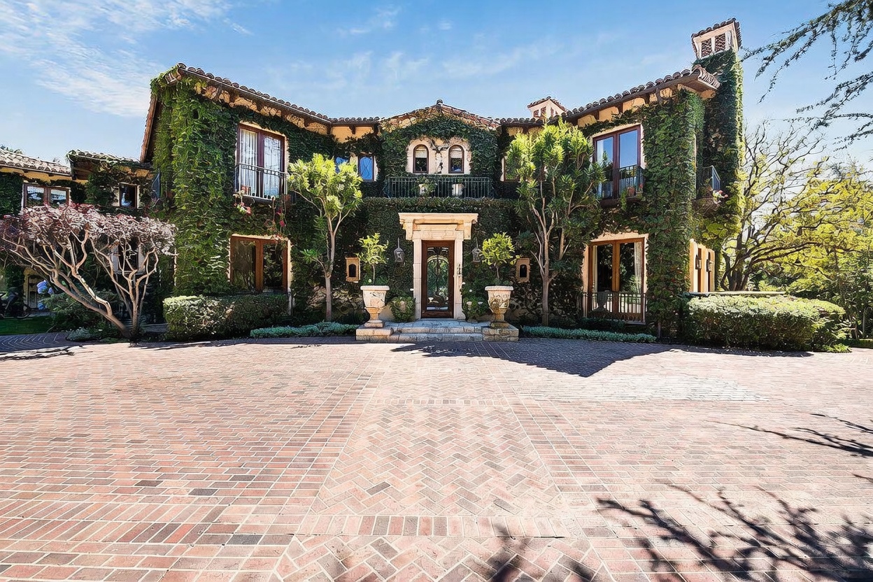This grand, Mediterranean-style estate features a striking facade completely covered in lush, climbing ivy. The front entrance is framed by a stone portico, leading to a large, circular brick-paved motor court that provides an expansive sense of arrival. The symmetrical design, arched windows, and mature landscaping create an atmosphere of timeless elegance and privacy.