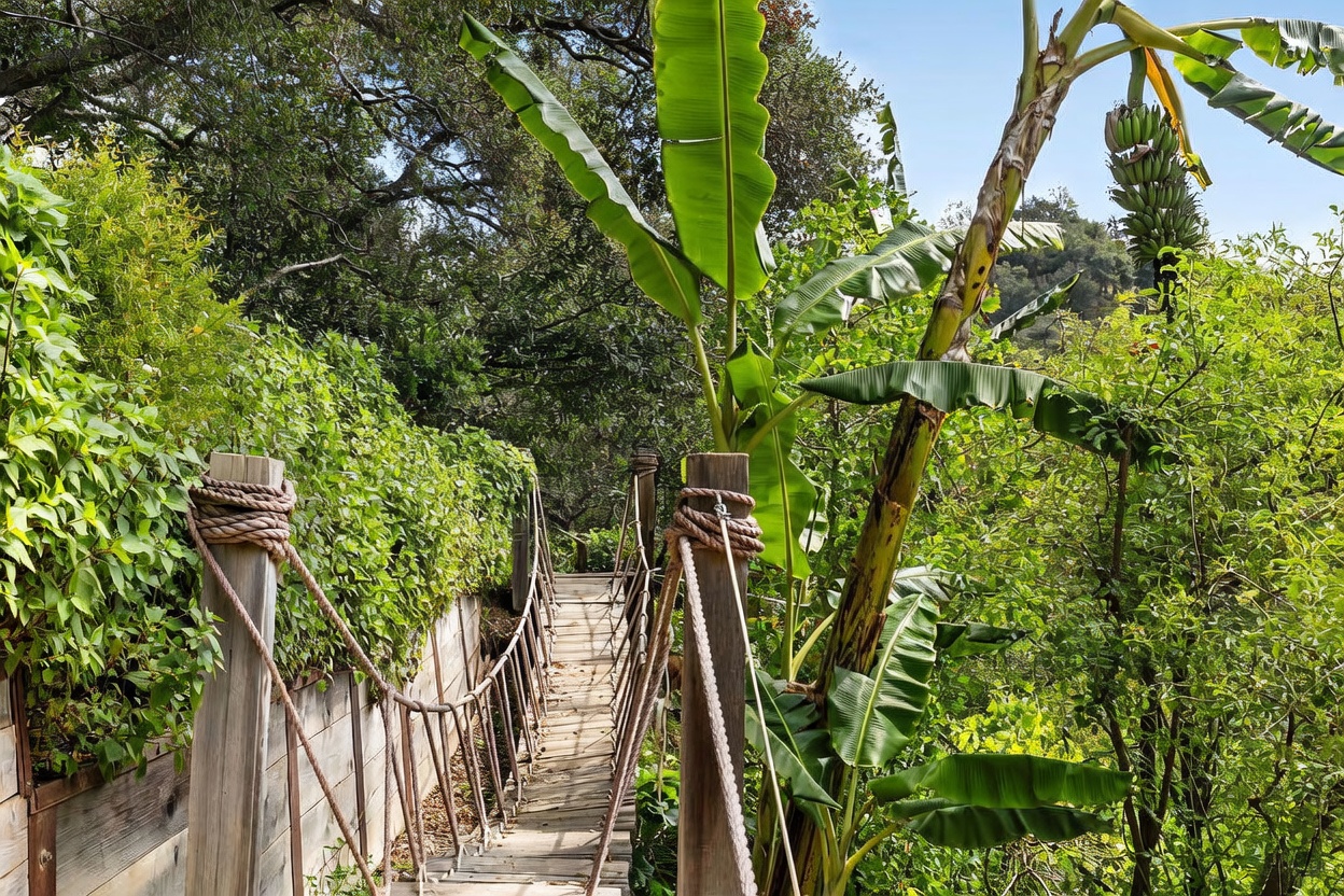 This image captures a rustic wooden suspension bridge winding through a lush, tropical-inspired garden setting. The bridge features thick rope railings secured to sturdy wooden posts, leading the eye into a dense canopy of mature trees and vibrant greenery, including a prominent banana plant with visible fruit. The perspective is from the entrance of the bridge, emphasizing a secluded, adventurous path that blends seamlessly into the natural landscape.