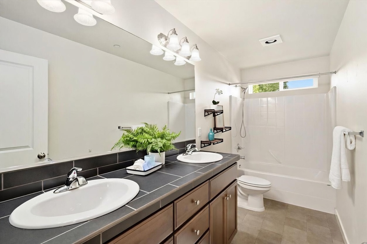 This bathroom features a dual-sink vanity with dark wood cabinetry and a dark tile countertop, complemented by a large wall-to-wall mirror. A shower-tub combination with white tile surround is positioned next to the toilet, and the room is finished with neutral-toned floor tiles. The perspective is from the doorway, capturing the functional layout and clean, well-lit atmosphere of the space.