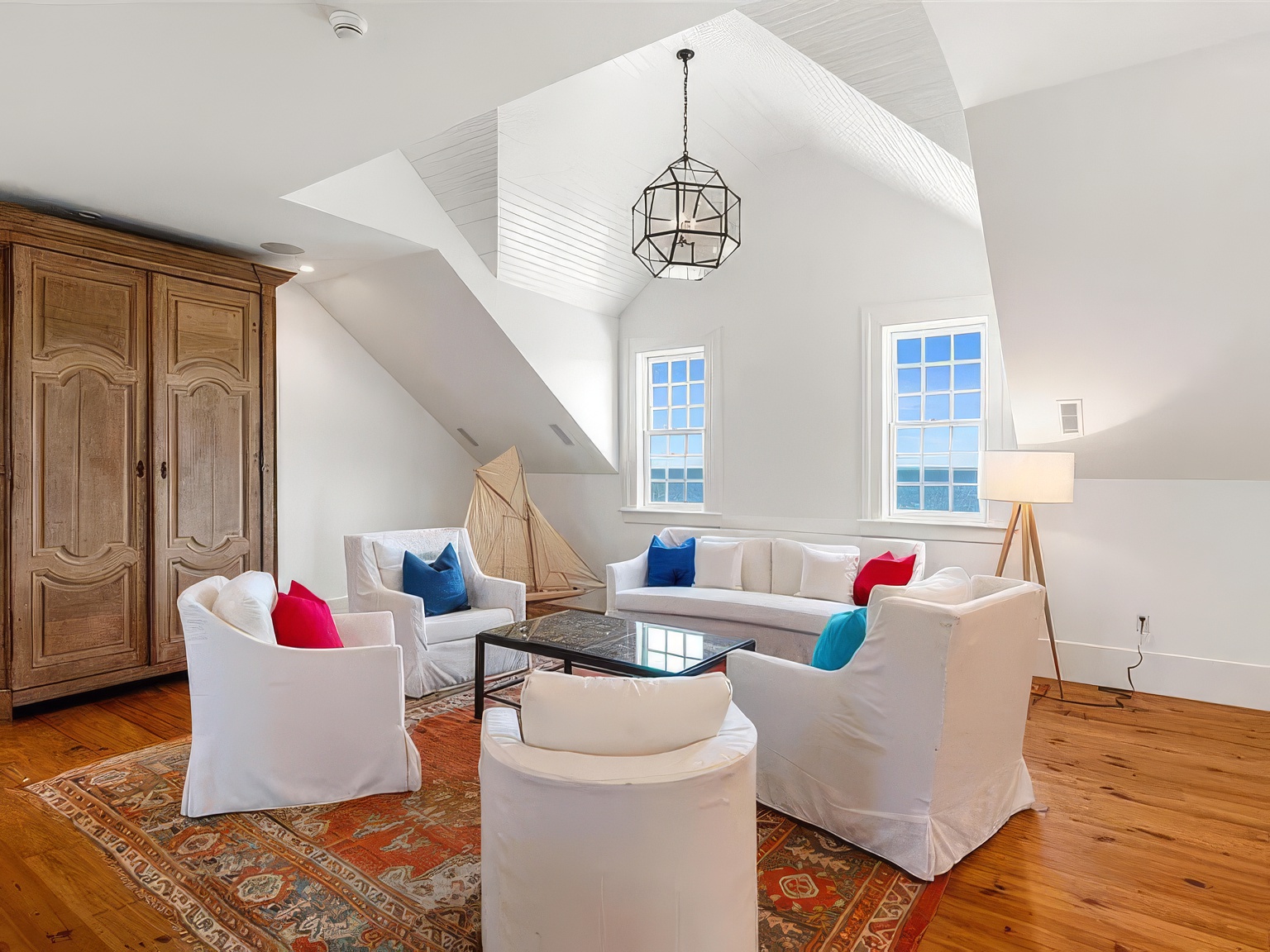 This bright and airy living room features a vaulted ceiling with white shiplap detailing and a prominent geometric pendant light. The space is furnished with white slipcovered seating arranged around a dark coffee table on a vibrant, patterned area rug, all set against warm wood flooring. Two large windows provide a clear view of the ocean, creating a serene and coastal-inspired atmosphere.
