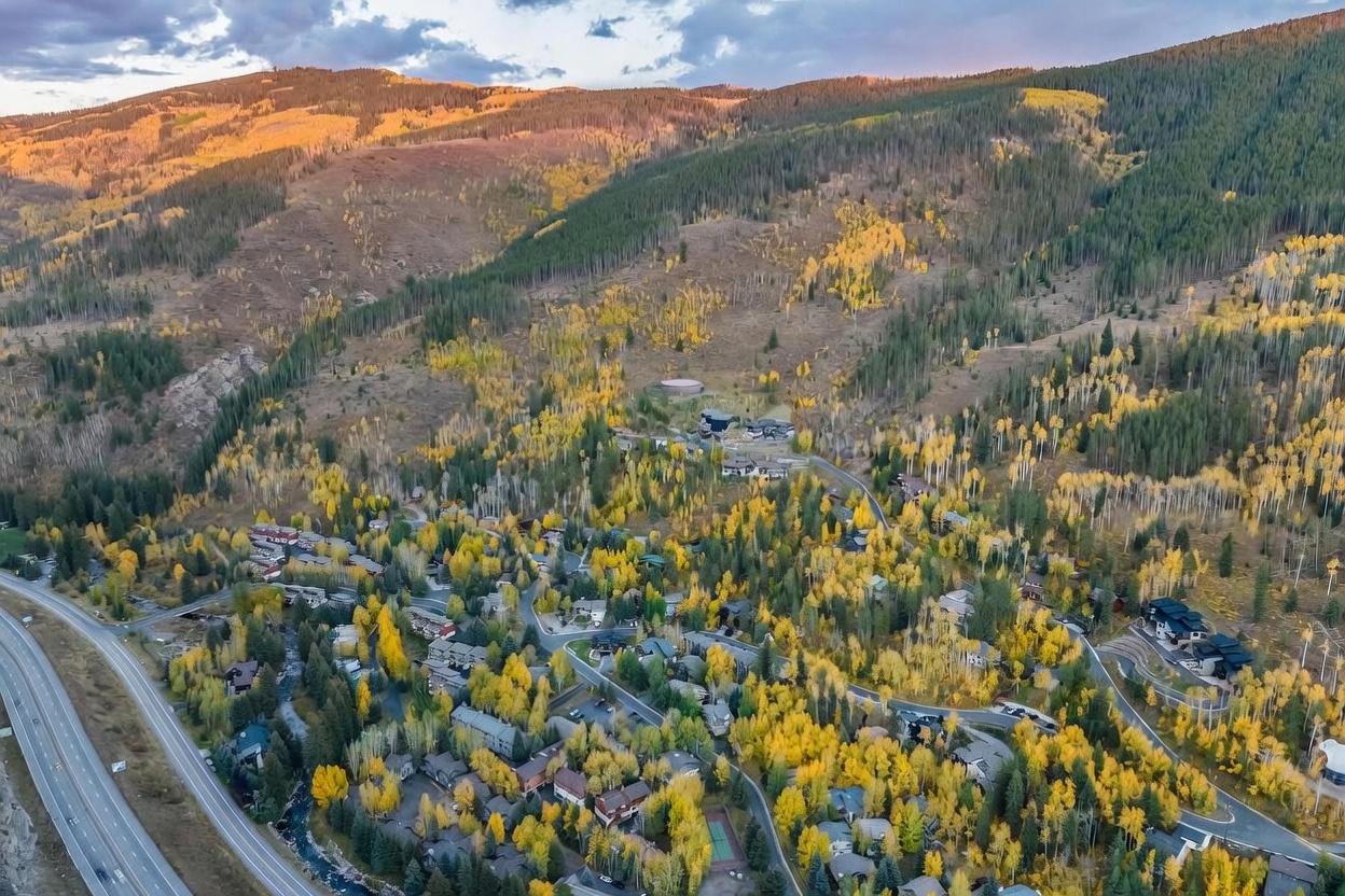 This high-altitude aerial perspective captures a sprawling mountain community nestled within a dense forest of golden-hued aspen trees during autumn. The scene features a network of winding roads connecting various residential properties, with a major highway running along the left edge of the frame. The dramatic backdrop of rugged, sun-drenched mountain slopes creates a serene and picturesque setting for this secluded neighborhood.