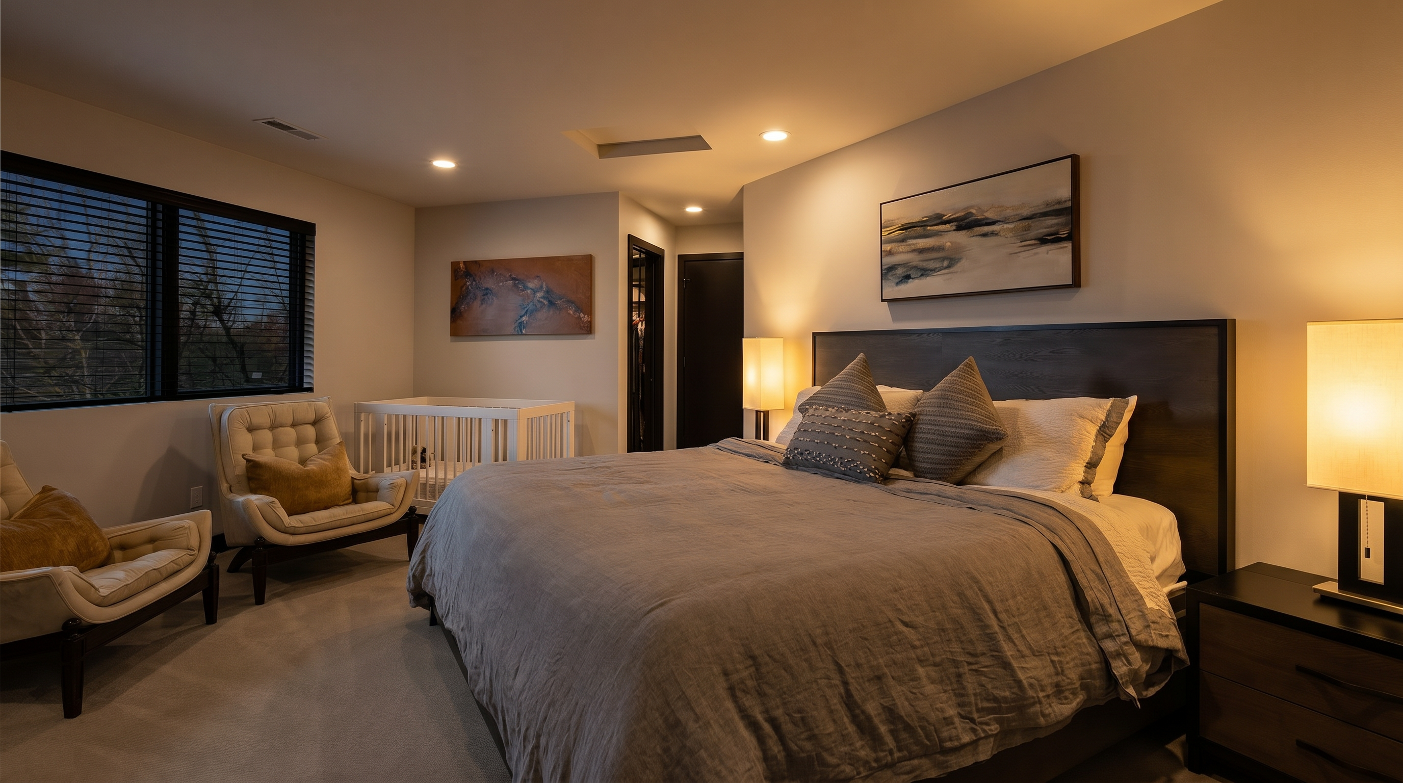 This spacious primary bedroom features a large bed with a dark wood headboard, neutral bedding, and two accent chairs positioned near a window. A baby crib is tucked into the corner, and the room is decorated with modern wall art, creating a serene and functional living space. The perspective is from the corner of the room, capturing the layout and the bright, airy atmosphere.