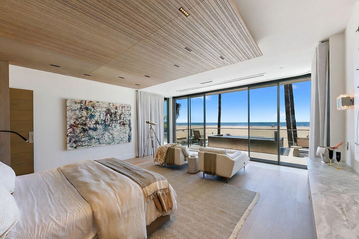 This luxurious primary bedroom features a sophisticated design with a slatted wood ceiling and light wood flooring. A large, abstract painting hangs above the bed, while floor-to-ceiling glass sliding doors provide an expansive, unobstructed view of the beach and ocean. The room is furnished with two cream-colored armchairs and a sleek stone-topped console, creating a serene and high-end coastal atmosphere.