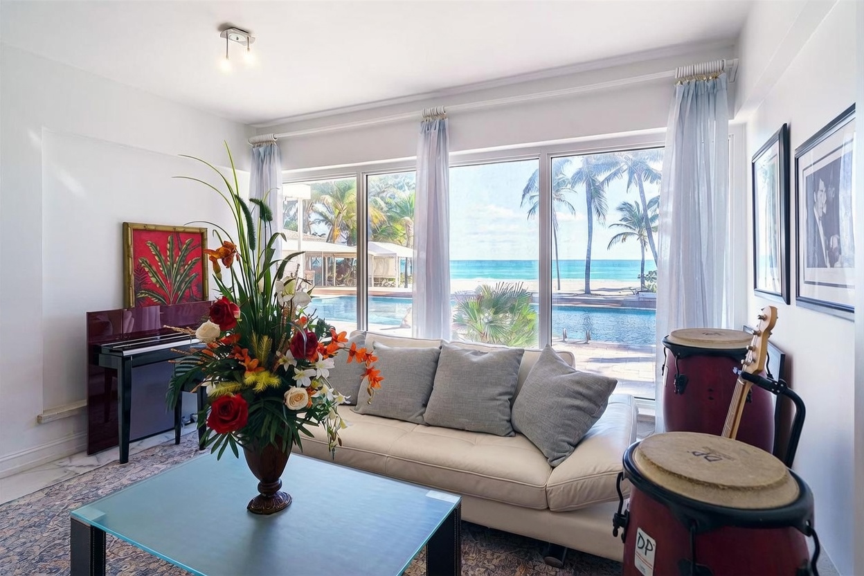 This bright living room features a cream-colored sofa positioned in front of large windows that offer a direct view of a swimming pool and the ocean beyond. The space is decorated with a large floral arrangement on a glass-topped coffee table, a piano, and musical drums, creating a relaxed, artistic atmosphere. The room is bathed in natural light, highlighting the neutral walls and the patterned area rug.