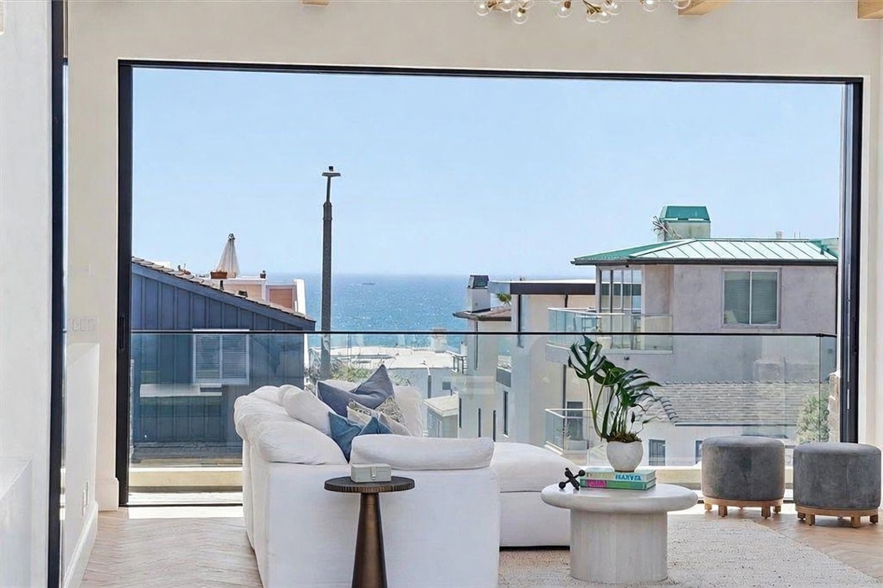 This bright living room features a large white sectional sofa positioned to take advantage of the expansive, floor-to-ceiling glass wall that frames a panoramic ocean view. The space is decorated with a neutral color palette, including a round stone-topped coffee table and two grey upholstered ottomans on a textured area rug. The herringbone-patterned light wood flooring adds a touch of warmth to the modern, airy atmosphere.