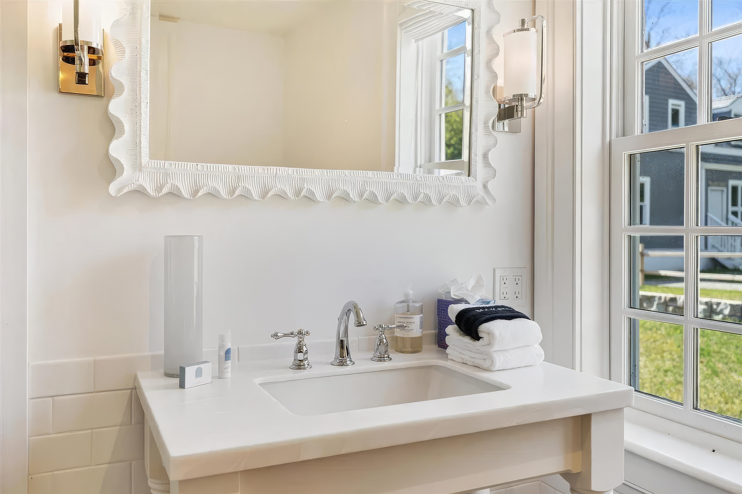 This bright bathroom vanity area features a white stone countertop with a classic undermount sink and polished chrome faucet fixtures. A decorative, textured-frame mirror hangs above the vanity, flanked by two wall-mounted sconces that provide soft lighting. To the right, a multi-pane window allows natural light to enter, while a stack of fresh white towels and a bottle of hand soap sit neatly on the counter.