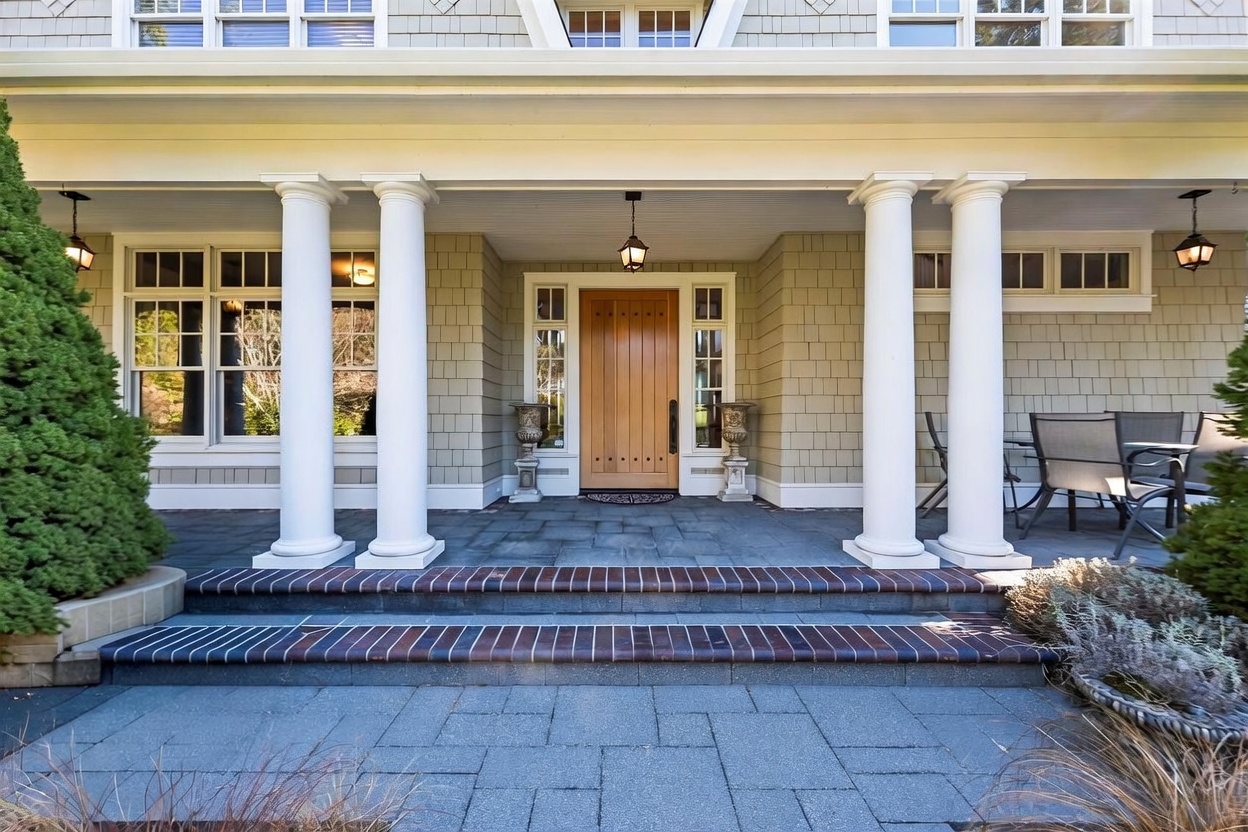 This inviting front entryway features a covered porch supported by four classic white columns, leading to a natural wood front door. The porch is paved with dark stone tiles and accented by two decorative urns, while the exterior walls are finished in light-colored shingles. A set of brick-edged steps provides a welcoming transition from the paved walkway to the porch area.