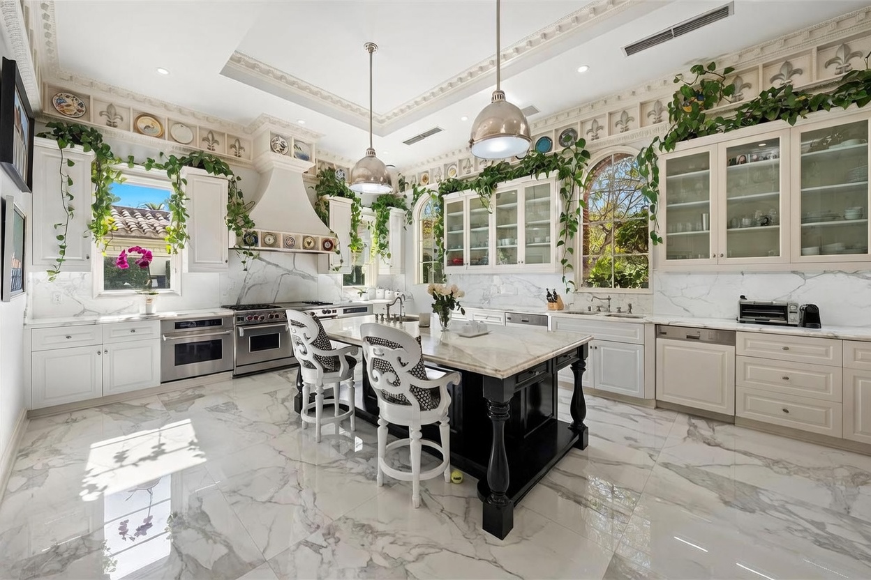 This elegant, high-end kitchen features a central island with a dark base and a light stone countertop, complemented by two ornate white bar stools. The space is framed by white cabinetry, light stone countertops, and a decorative range hood, all accented by lush greenery trailing along the upper wall molding. Large arched windows allow natural light to flood the room, highlighting the polished stone flooring and the sophisticated, classic aesthetic of the space.