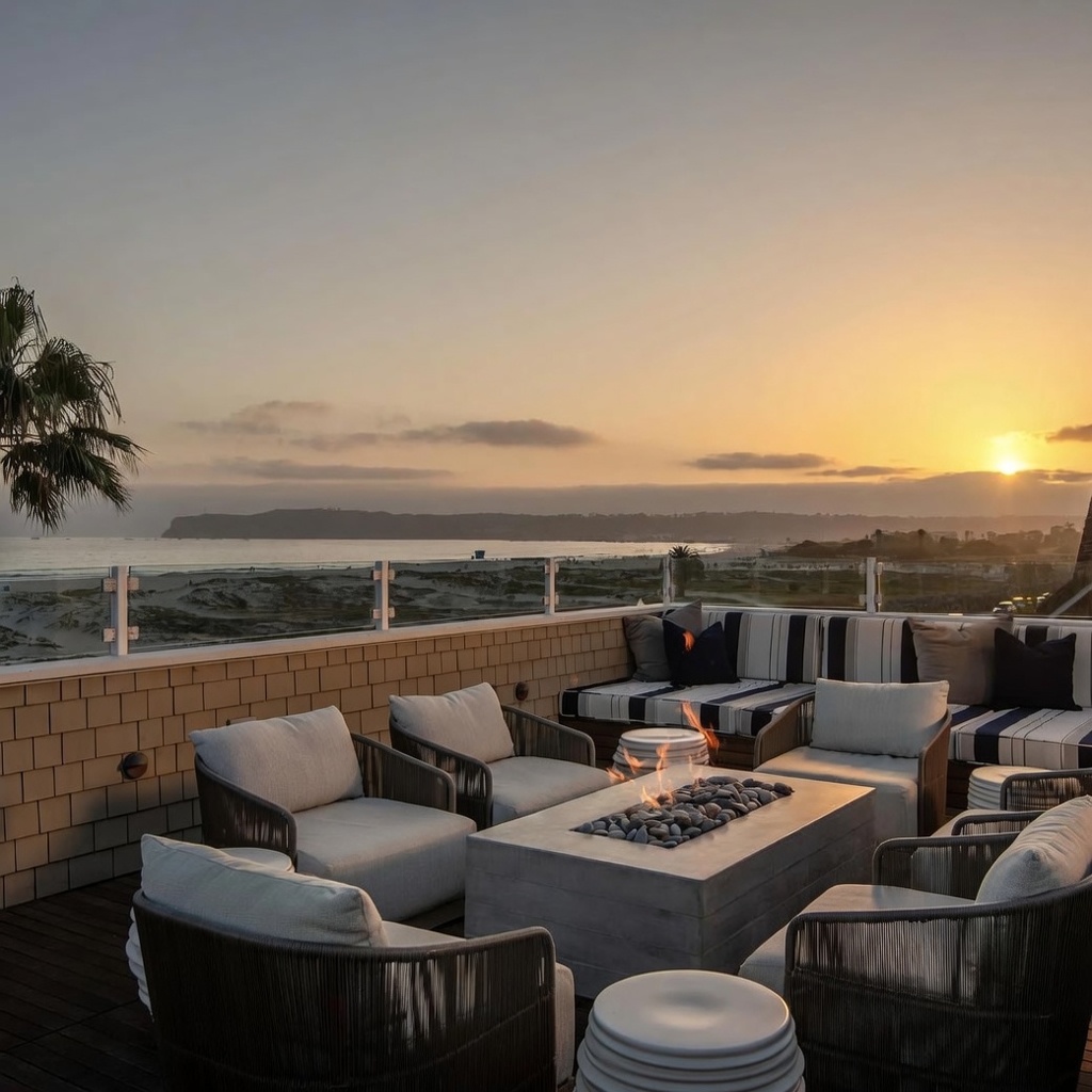 This expansive rooftop deck offers a luxurious outdoor living space with a panoramic view of the ocean and coastline at sunset. The area is furnished with comfortable lounge chairs and a built-in bench, all centered around a rectangular concrete fire pit filled with smooth stones. The setting provides a serene and sophisticated atmosphere, perfect for evening relaxation while overlooking the beach.