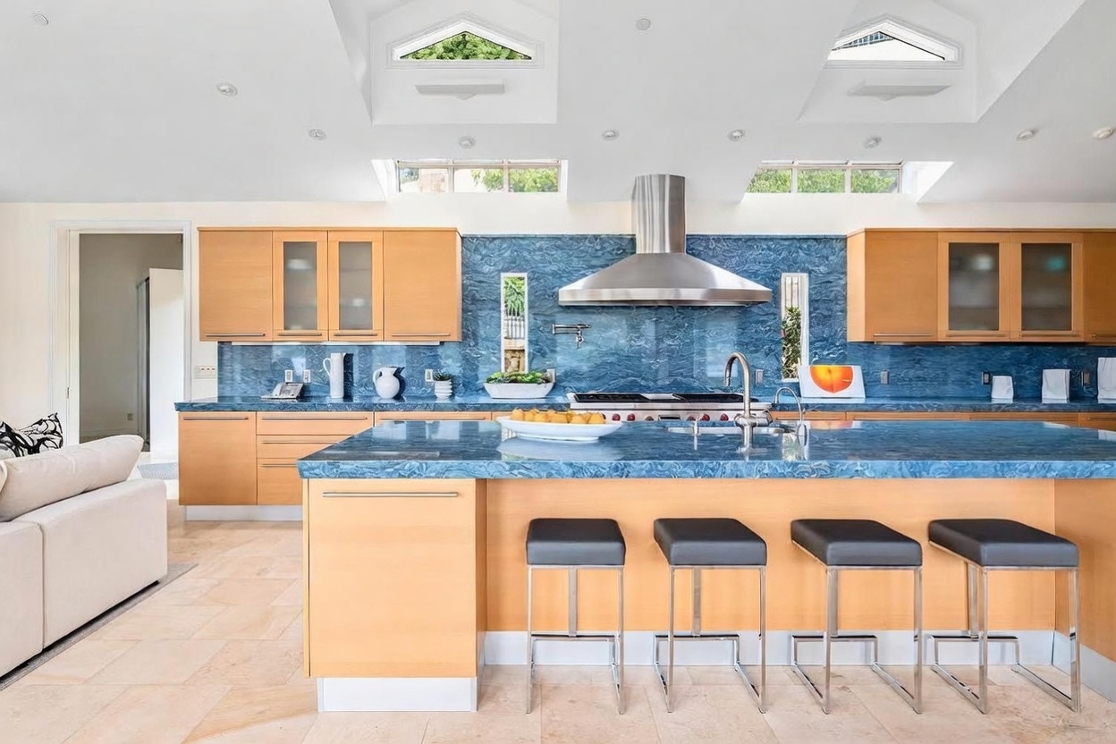 This bright and airy kitchen features a striking blue stone countertop and matching backsplash that contrast with the light wood cabinetry. The space is illuminated by multiple skylights and recessed lighting, highlighting the large central island with bar seating. The perspective is a wide-angle shot from the adjacent living area, showcasing an open-concept layout perfect for entertaining.