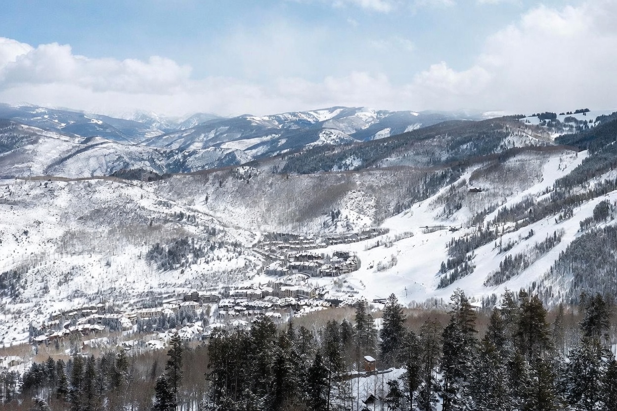 This expansive aerial view captures a picturesque mountain resort nestled within a snow-covered valley. The scene features a dense cluster of residential buildings and lodges surrounded by groomed ski runs and towering, evergreen-forested slopes under a bright, cloudy sky. The perspective provides a grand, cinematic overview of the community's integration into the rugged alpine landscape.
