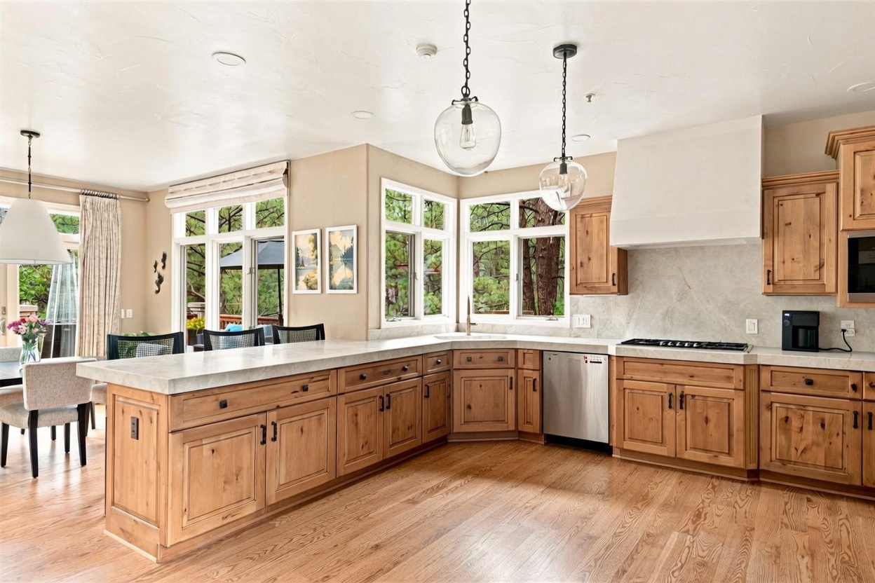 This spacious kitchen features warm-toned wood cabinetry paired with light-colored stone countertops and a matching backsplash. A large central island provides additional workspace and built-in shelving for books, while two glass pendant lights hang above to illuminate the area. Large windows offer a scenic view of the surrounding trees, creating a bright and inviting atmosphere.