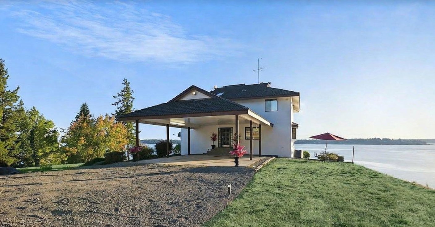 This waterfront property features a prominent covered carport leading to the main entrance, set against a backdrop of a calm bay and clear blue sky. The exterior is finished in a light-colored stucco, complemented by a dark-tiled roof and a gravel driveway that transitions into a manicured lawn. The perspective is a wide, eye-level shot that captures the home's integration with the surrounding natural landscape and water views.