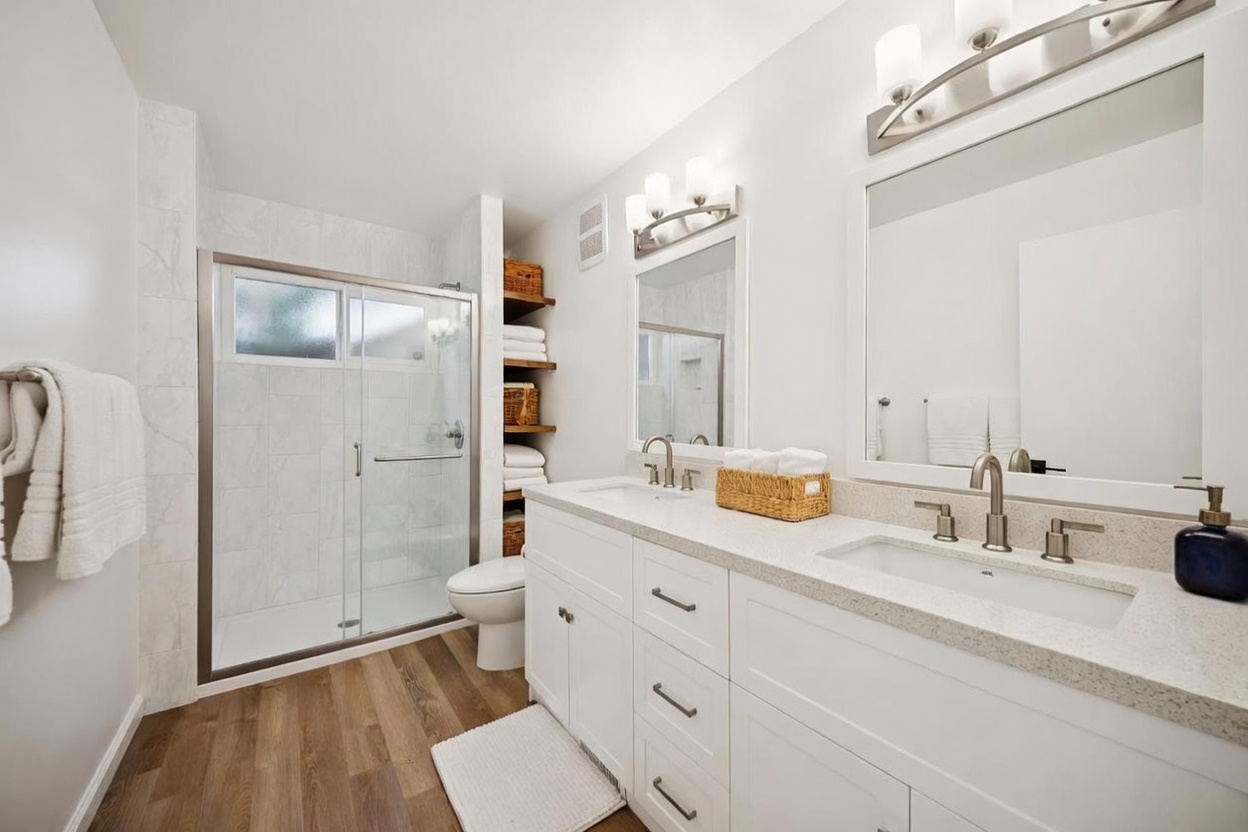 This bright and modern bathroom features a double vanity with white cabinetry and a light-toned stone countertop. To the left, a glass-enclosed shower with light-colored tile walls sits adjacent to a toilet and open shelving for storage. The room is finished with wood-look flooring and dual mirrors illuminated by contemporary light fixtures, creating a clean and functional aesthetic.