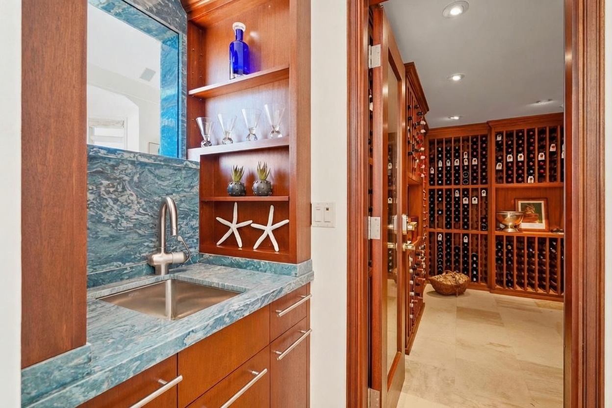 This image captures a sophisticated wet bar area adjacent to a climate-controlled wine cellar. The bar features a striking blue-toned stone countertop and backsplash, complemented by warm wood cabinetry and open shelving decorated with glassware and coastal accents. Through the glass-paneled door, the wine cellar is visible, showcasing extensive wooden racking systems and a neutral-toned stone floor, creating an elegant space for entertaining.