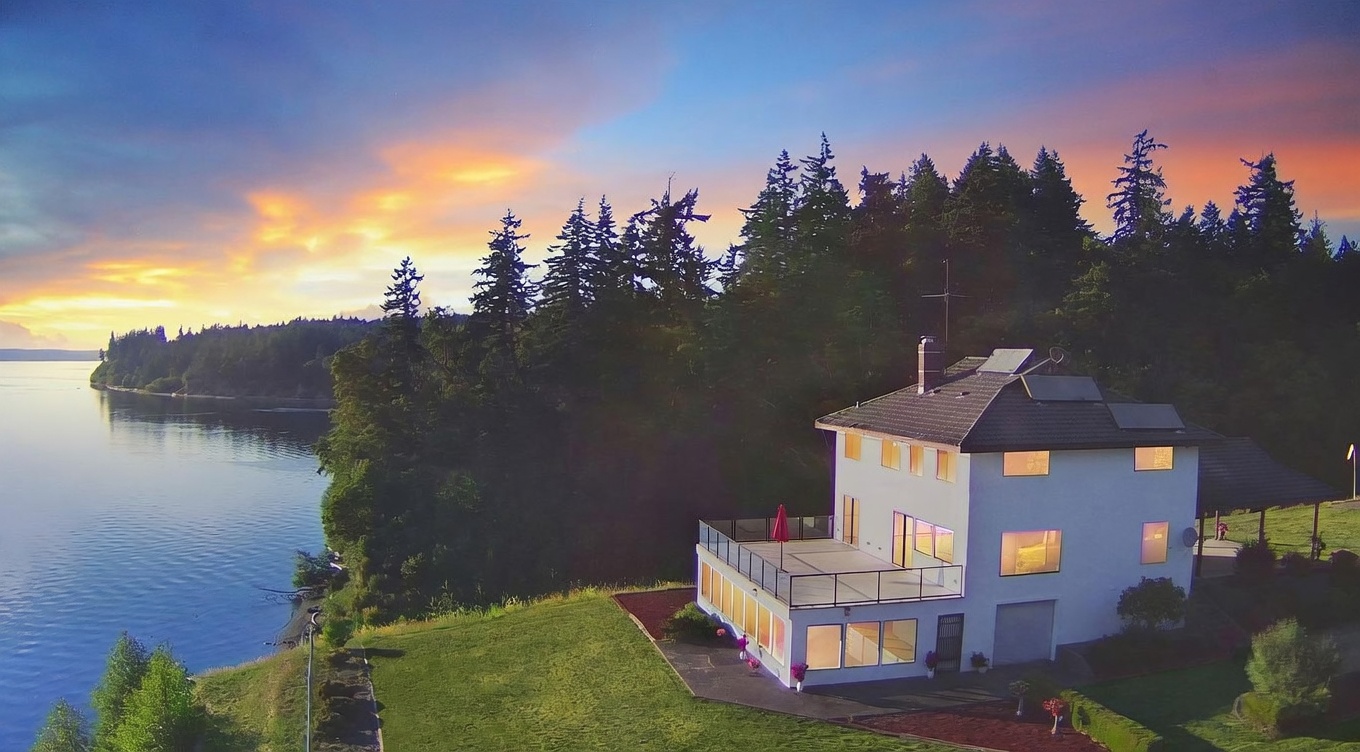 This aerial perspective captures a multi-level home perched on a grassy bluff overlooking a serene body of water at sunset. The house features a prominent upper-level deck with glass railings, large windows, and a light-colored exterior, set against a backdrop of dense evergreen trees. The composition emphasizes the property's expansive waterfront location and tranquil, scenic setting.