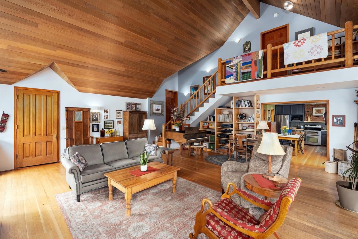 This spacious, open-concept living room features a dramatic vaulted wood-paneled ceiling and a cozy, rustic aesthetic. The room is furnished with a comfortable grey sofa, a wooden coffee table, and a patterned armchair, all anchored by a large area rug on light wood flooring. A staircase leads to an upper loft area, while a piano and built-in shelving units add character to the space, which flows directly into a bright kitchen in the background.