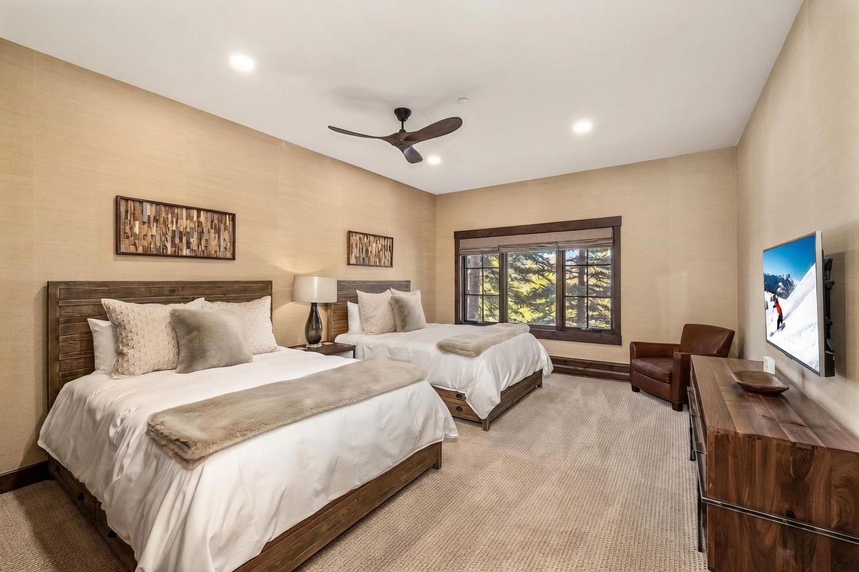 This spacious guest bedroom features two queen-sized beds with rustic wood headboards and neutral-toned bedding. The room is accented by textured wall coverings, a leather armchair near the window, and a wooden dresser supporting a flat-screen television. Large windows provide natural light and a view of the surrounding trees, creating a warm and inviting mountain-style retreat.