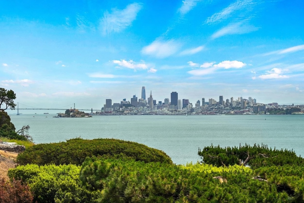 This scenic landscape view captures the San Francisco skyline across the bay, featuring Alcatraz Island in the mid-ground and the Bay Bridge extending to the left. The foreground is filled with lush, vibrant green pine trees and shrubbery, providing a natural frame for the iconic urban vista. The bright blue sky with wispy clouds creates a serene and expansive atmosphere, highlighting the property's prime location and panoramic outlook.