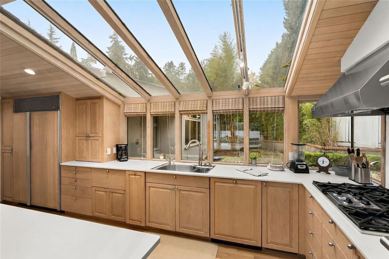 This bright, airy kitchen features light wood cabinetry and a long, white countertop that runs beneath a dramatic sloped glass ceiling. The space is filled with natural light, offering a clear view of the lush greenery outside through a series of windows. A stainless steel sink, a gas cooktop, and various small appliances are integrated into the functional layout, creating a warm and inviting atmosphere.